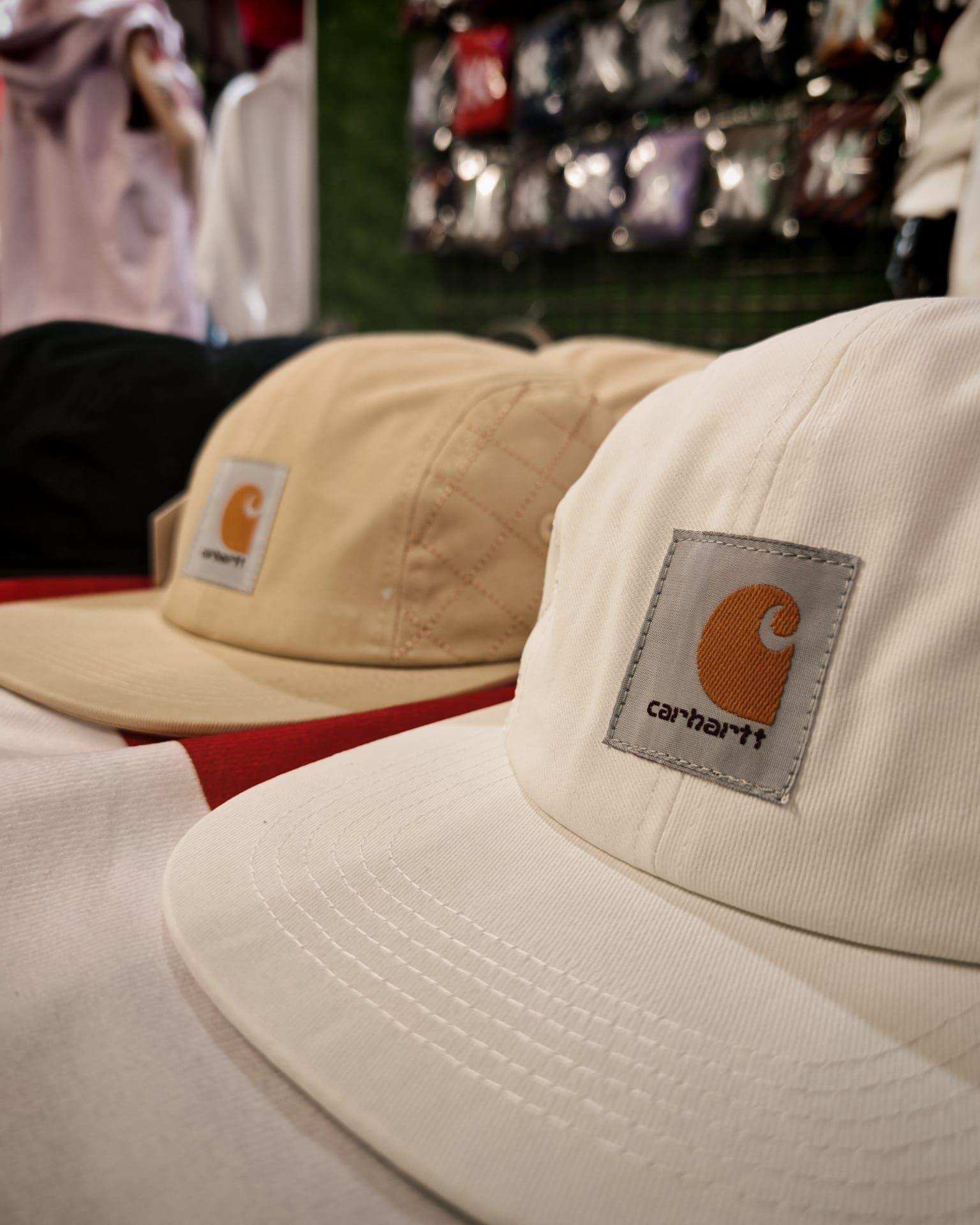 Due cappellini Carhartt beige in esposizione in un negozio, uno con la visiera rossa sul davanti