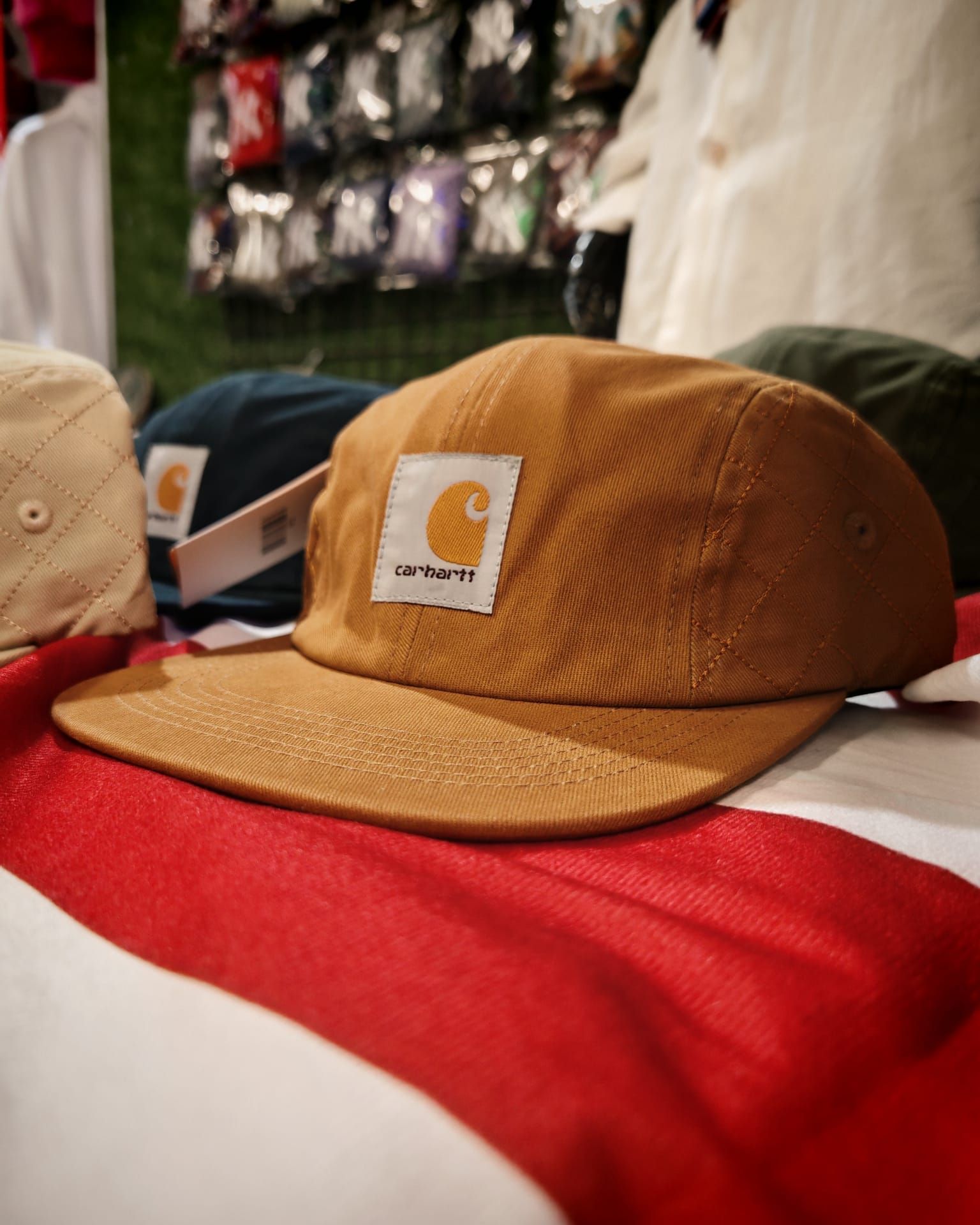 Un berretto Carhartt color cachi su una superficie a strisce rosse e bianche, con un secondo berretto sfocato sullo sfondo.