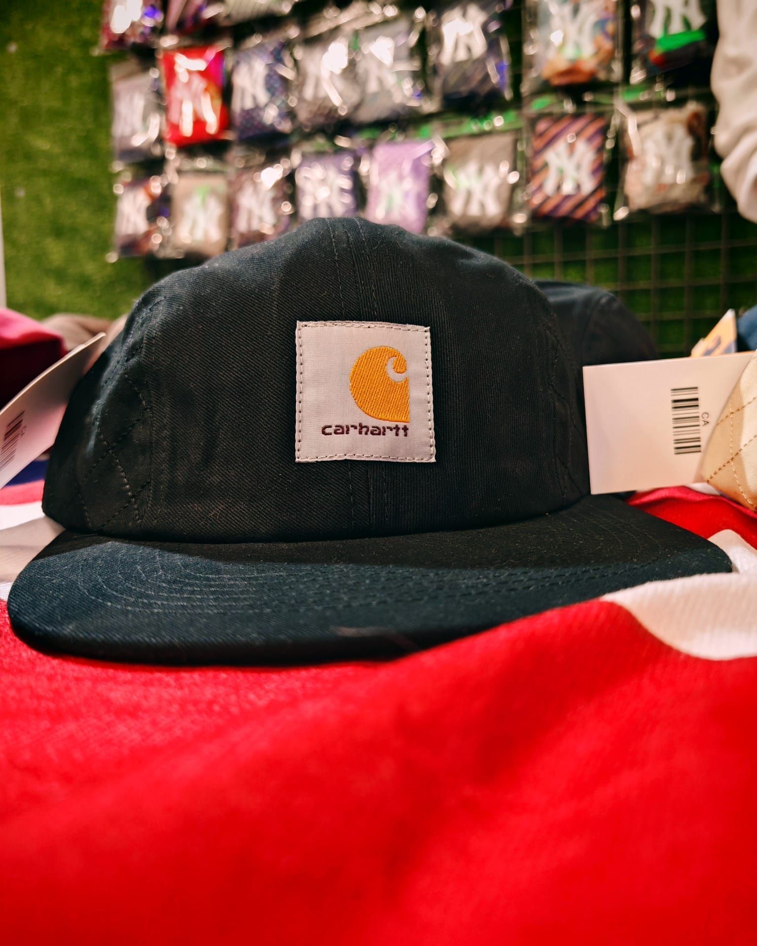 Berretto nero Carhartt su una superficie rossa, con accessori appesi sullo sfondo.