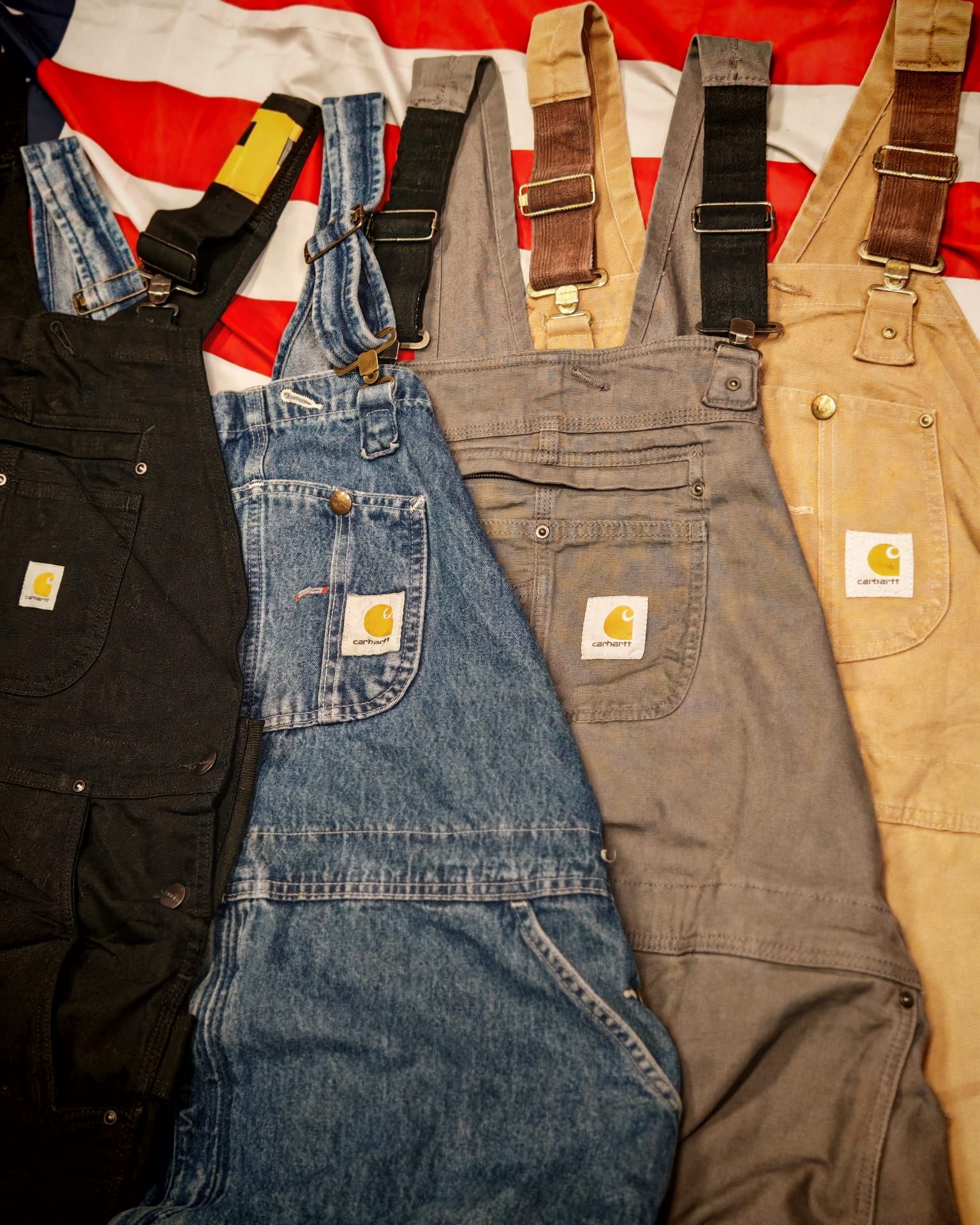 Quattro paia di salopette in stile Carhartt, nei colori nero, blu denim, grigio e beige, disposte una accanto all'altra.