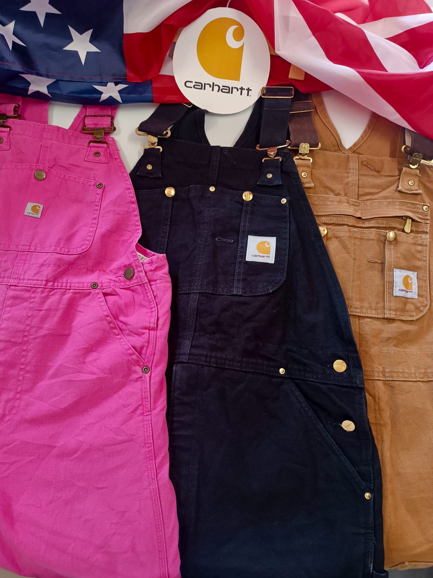 Tre salopette Carhartt rosa, nere e beige disposte su uno sfondo con la bandiera americana.