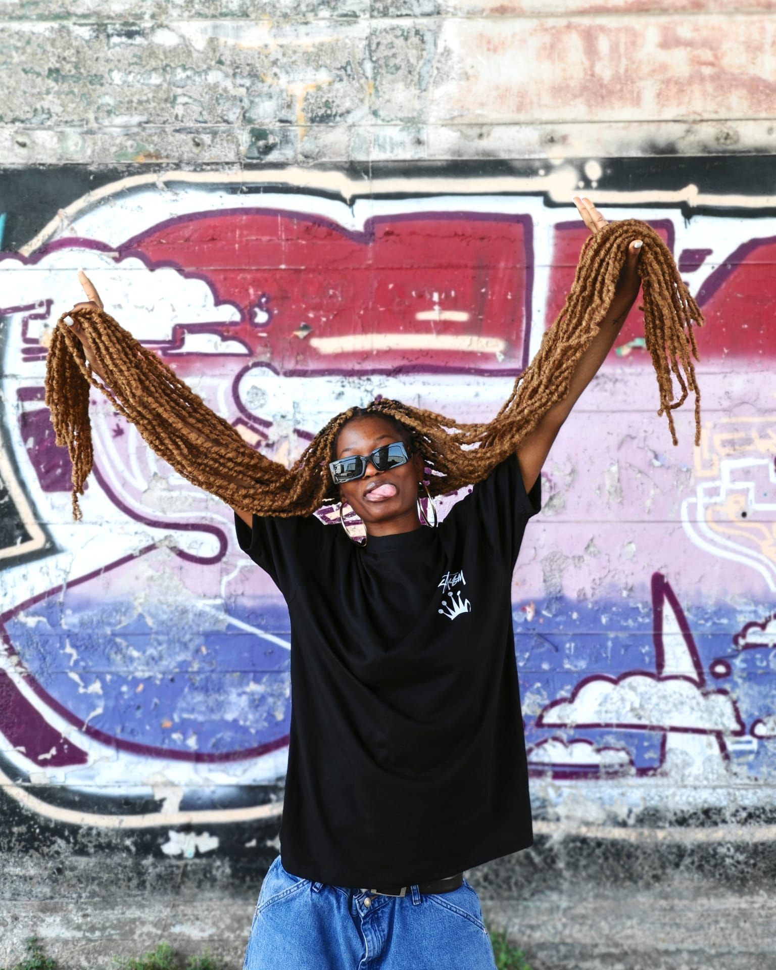 Persona con occhiali da sole in posa con le braccia alzate davanti a un muro ricoperto di graffiti colorati.
