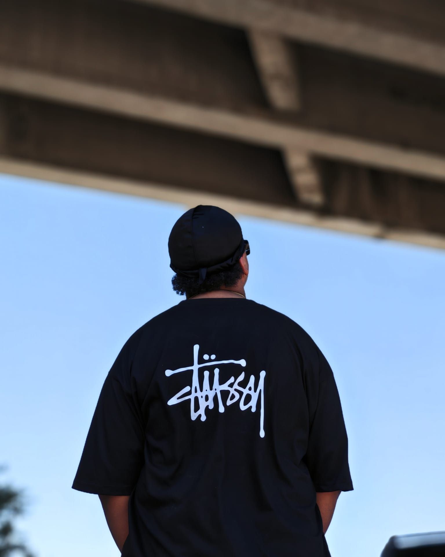 Persona con indosso una maglietta nera di Stüssy, vista di spalle sotto un cavalcavia di cemento, contro un cielo azzurro.
