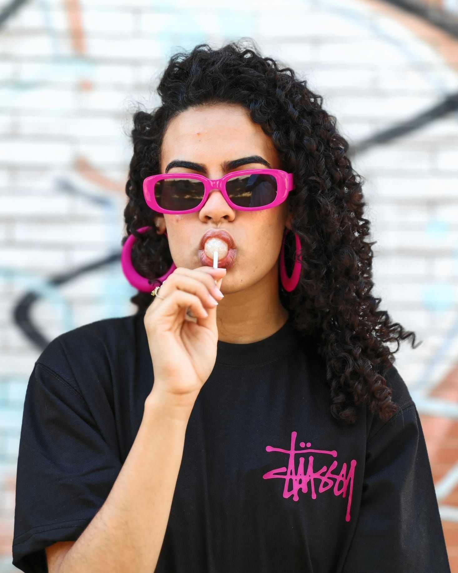 Una persona con occhiali da sole rosa mangia un lecca-lecca, indossando una maglietta nera, davanti a un muro ricoperto di graffiti.