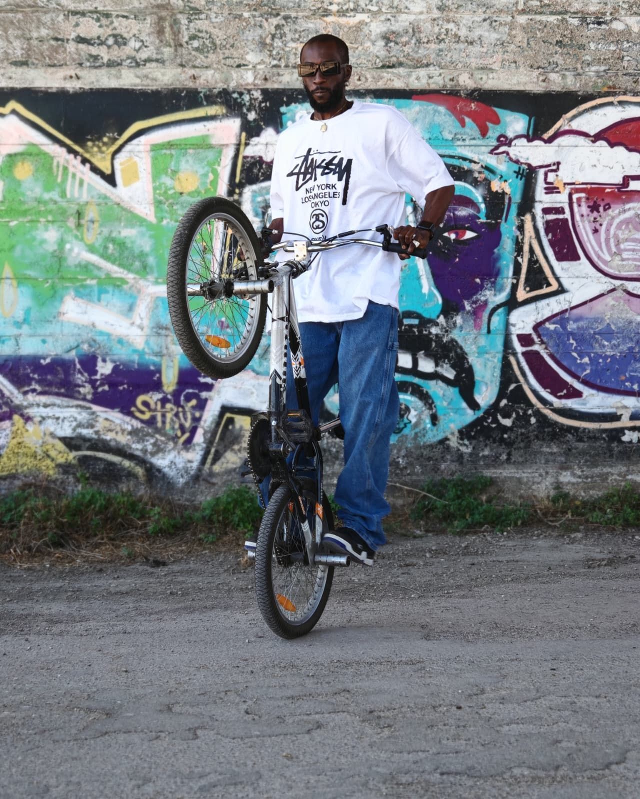 Un uomo in equilibrio su una bicicletta, su una sola ruota, davanti a un muro ricoperto di graffiti colorati.