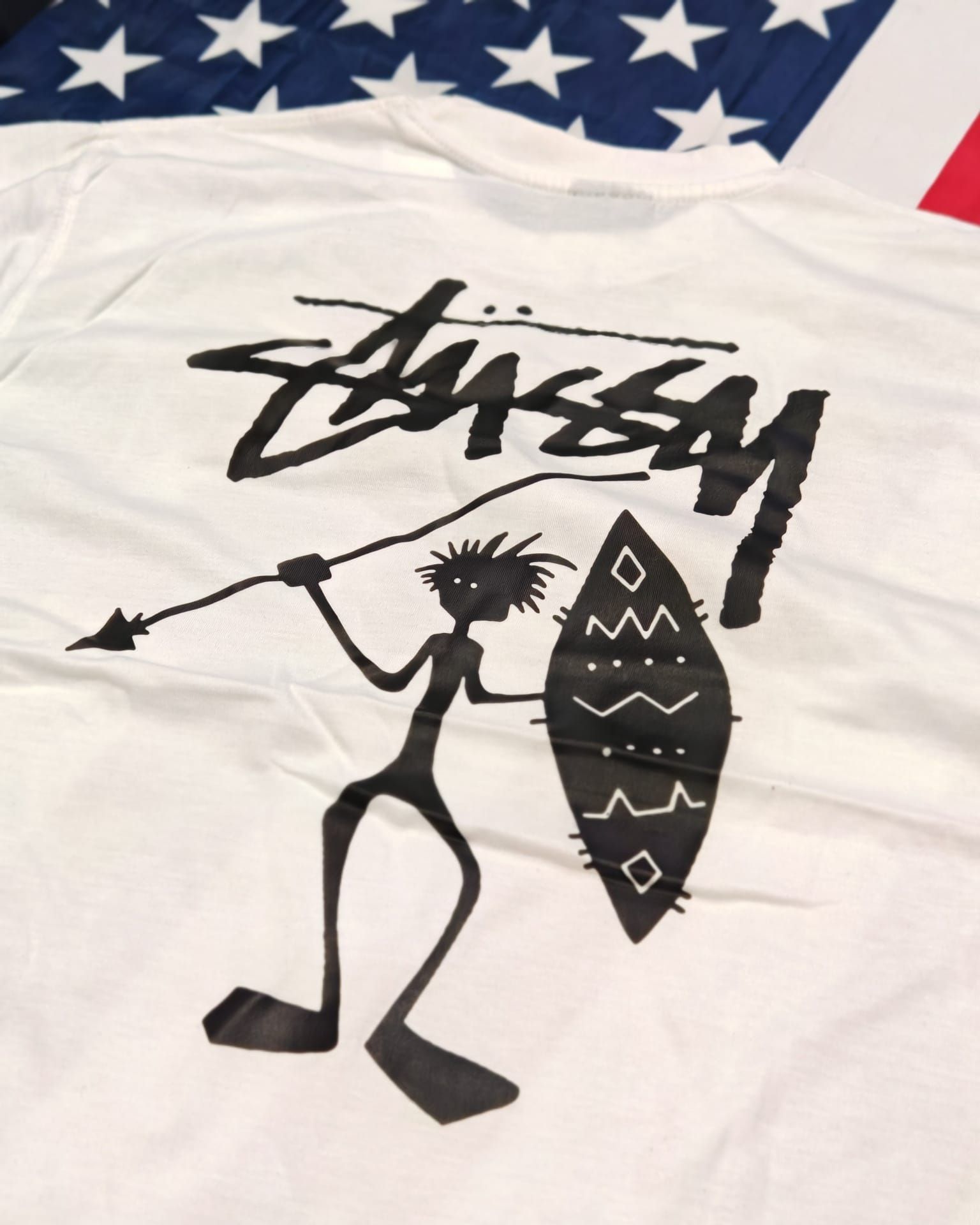 T-shirt bianca Stüssy con logo nero e omino stilizzato che tiene una tavola da surf; sullo sfondo si intravede la bandiera americana.