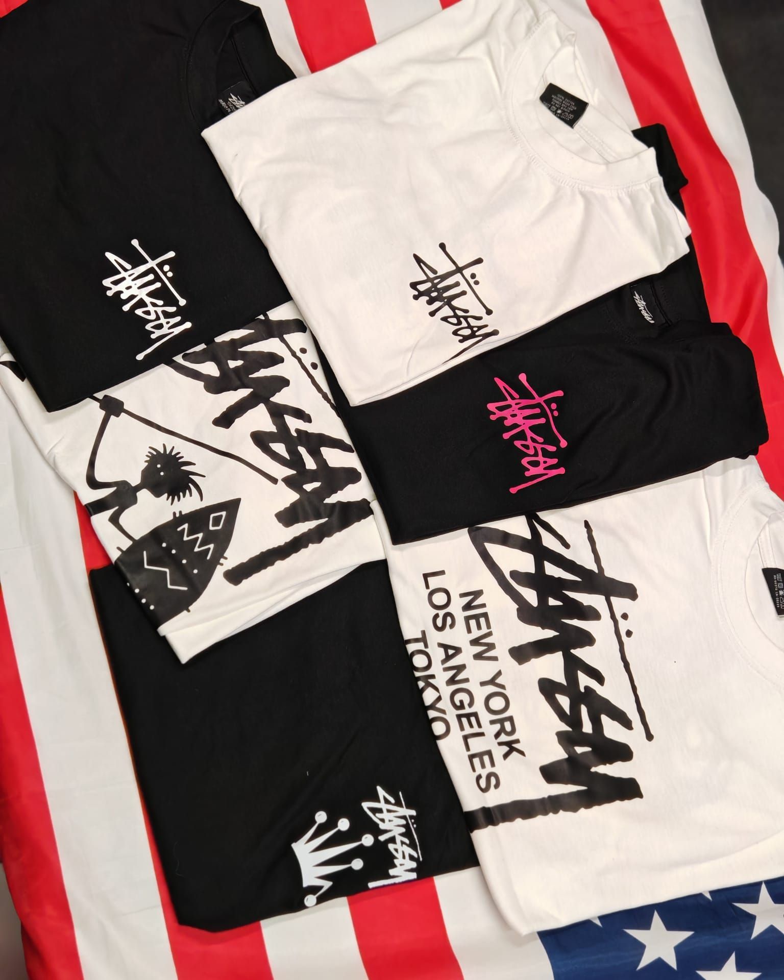Diverse t-shirt grafiche Stüssy in bianco e nero disposte su una superficie a strisce rosse, bianche e blu.