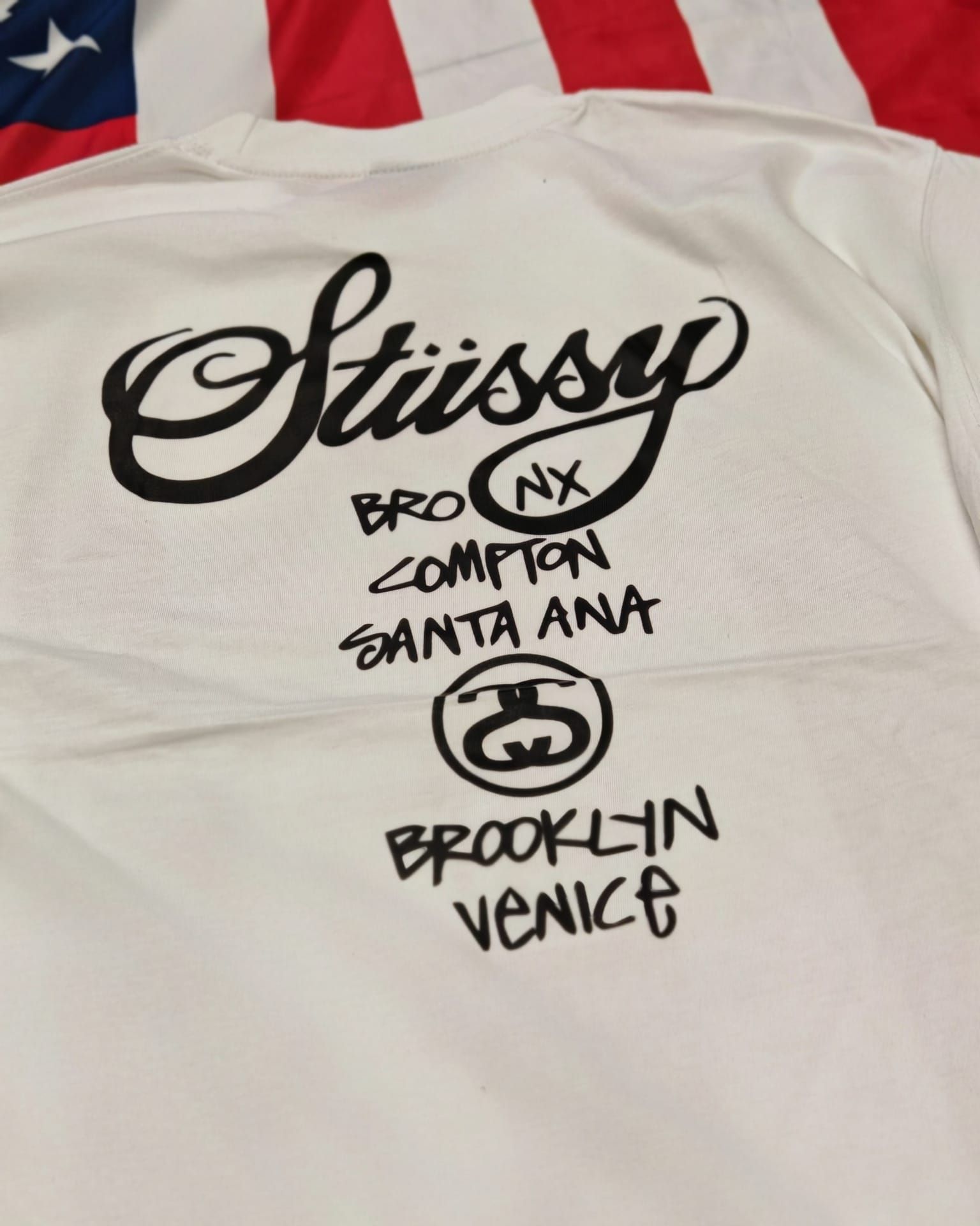 Maglietta bianca Stüssy con scritta nera in stile graffiti: Bronx, Compton, Santa Ana, Brooklyn, Venice.