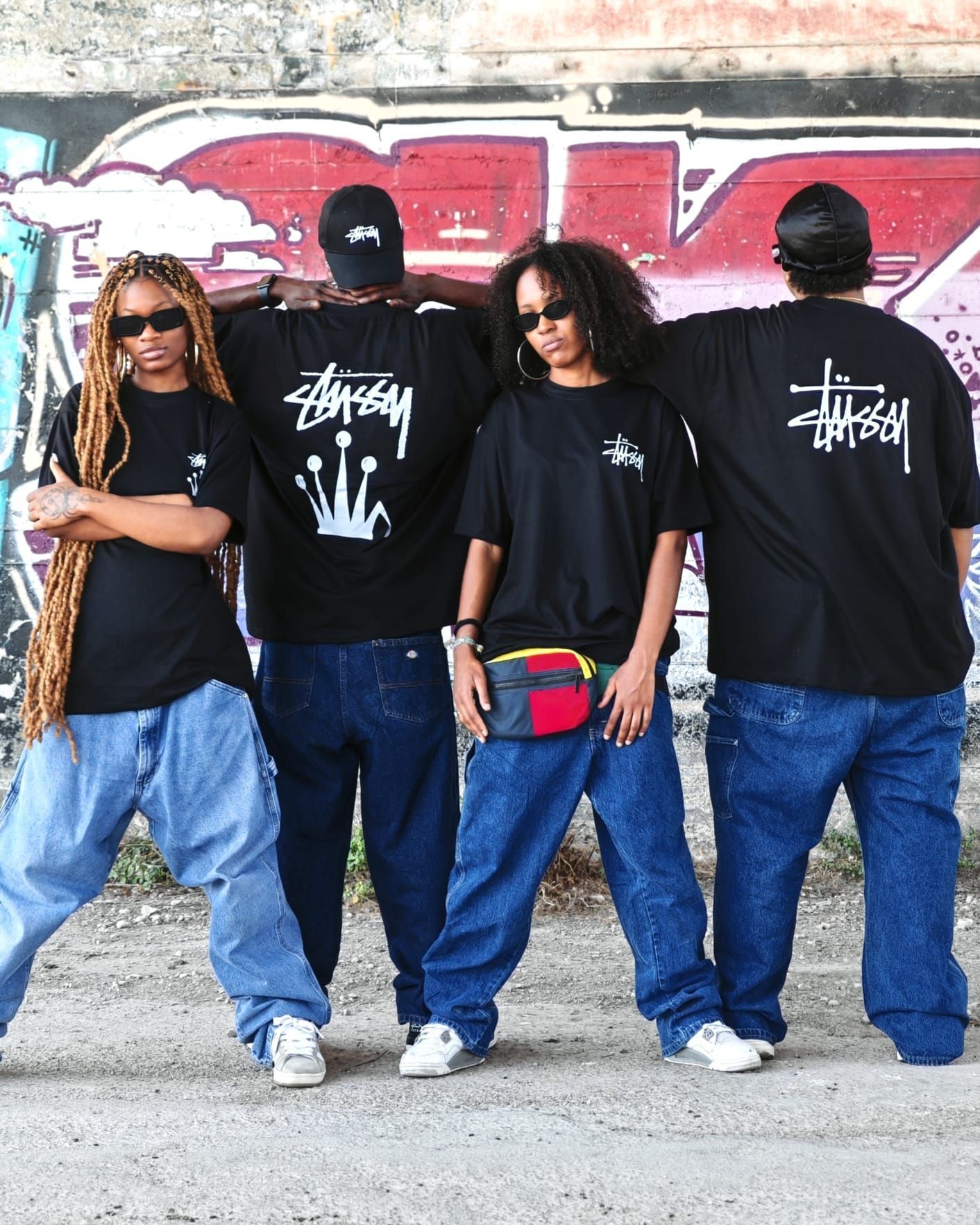 Quattro persone con magliette nere e jeans larghi in posa davanti a un muro ricoperto di graffiti.