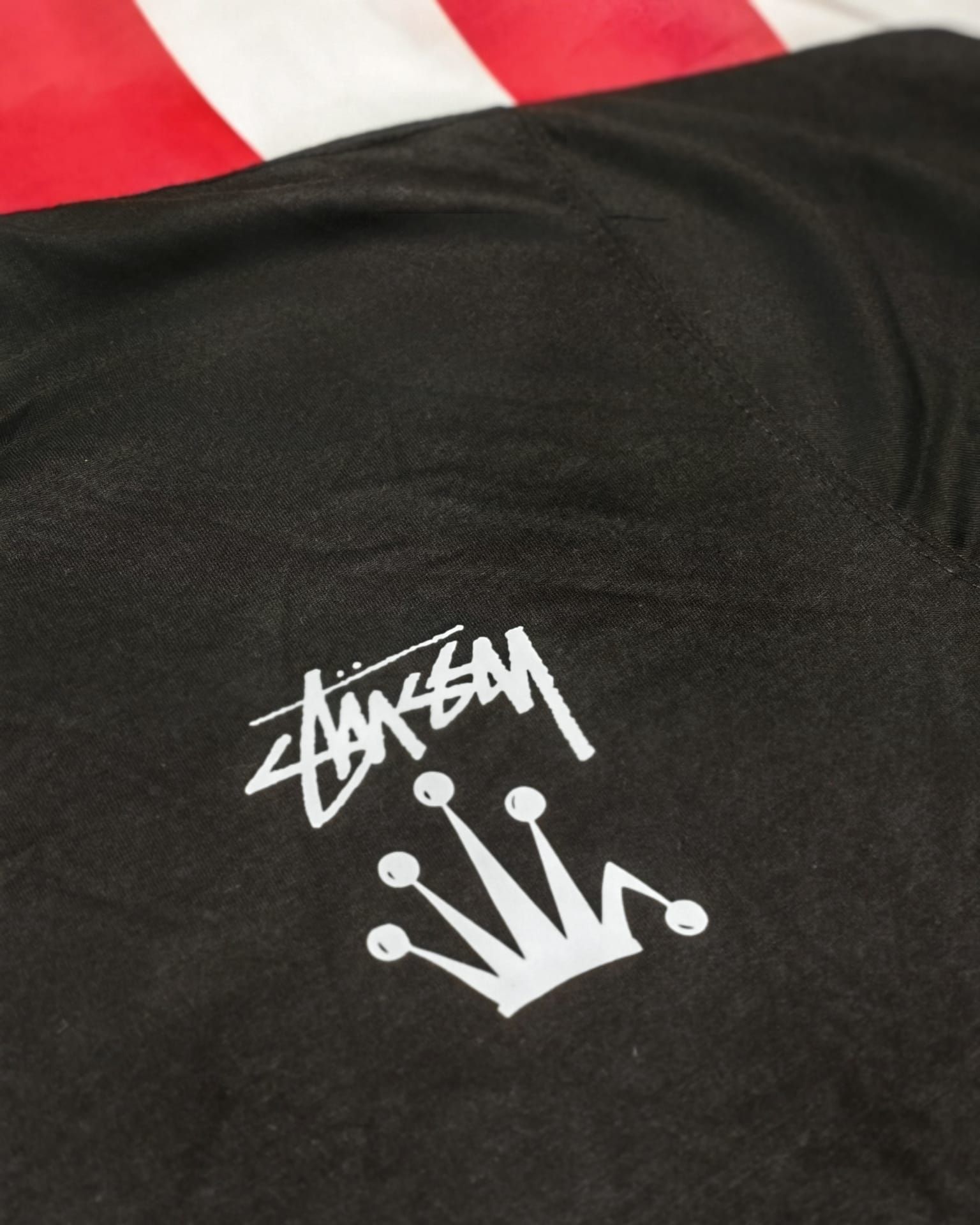Maglietta nera con logo Stüssy nero e grafica a corona bianca, sullo sfondo tessuto rosso e bianco.