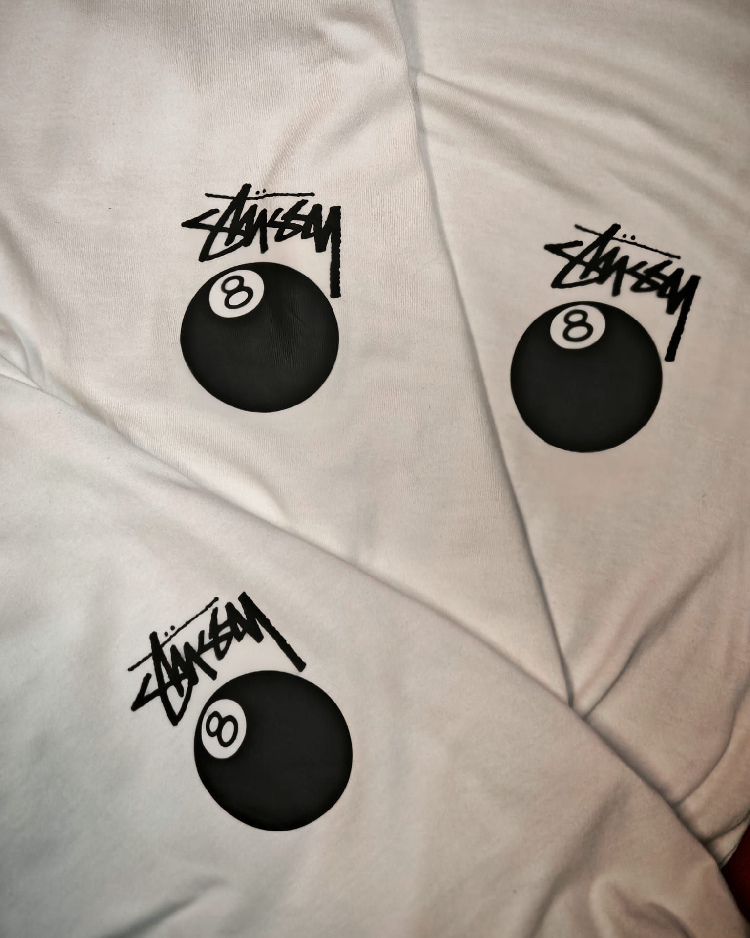 Primo piano di tre grafiche nere a forma di palla con il logo Stüssy su uno sfondo di tessuto chiaro.