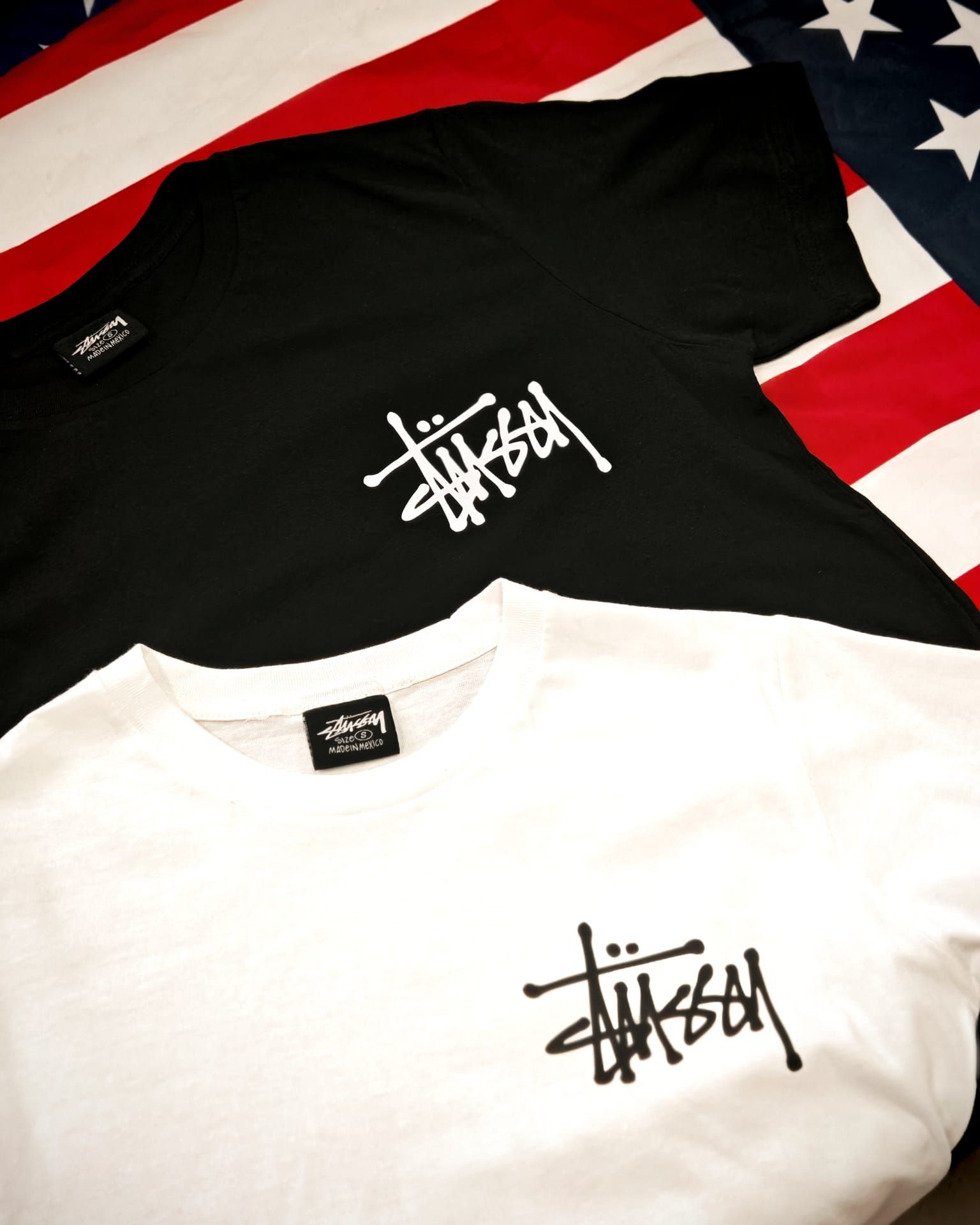 Due t-shirt con il logo Stüssy, una nera e una bianca, adagiate su uno sfondo con la bandiera americana.
