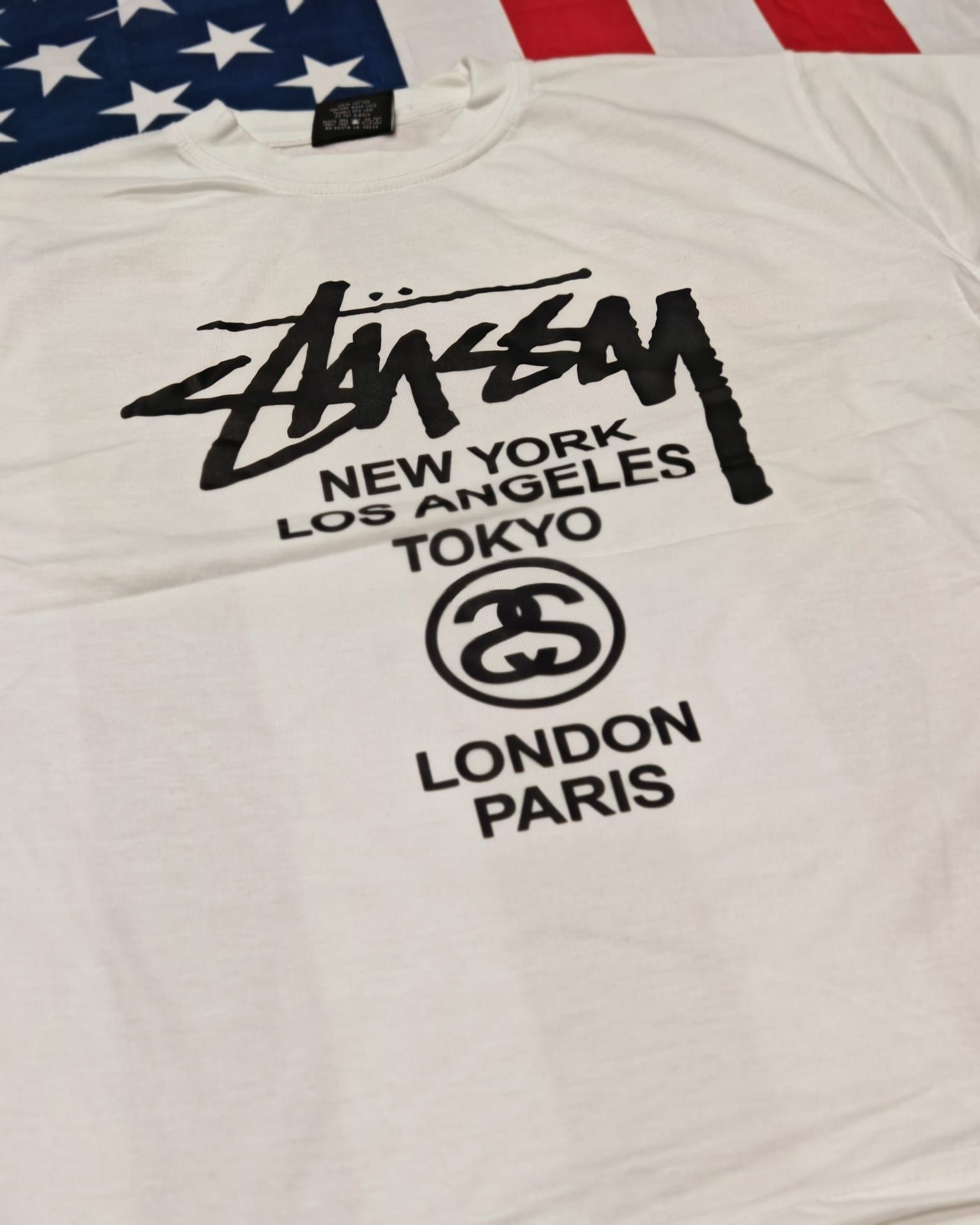T-shirt bianca Stüssy con logo nero e nomi delle città: New York, Los Angeles, Tokyo, Londra, Parigi