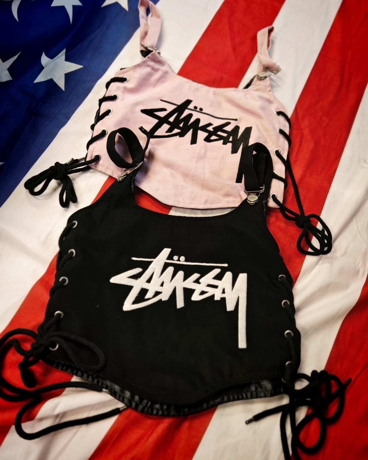 Tre borse a sacco Stussy con coulisse, nei colori nero, bianco e rosa, su uno sfondo con la bandiera americana.