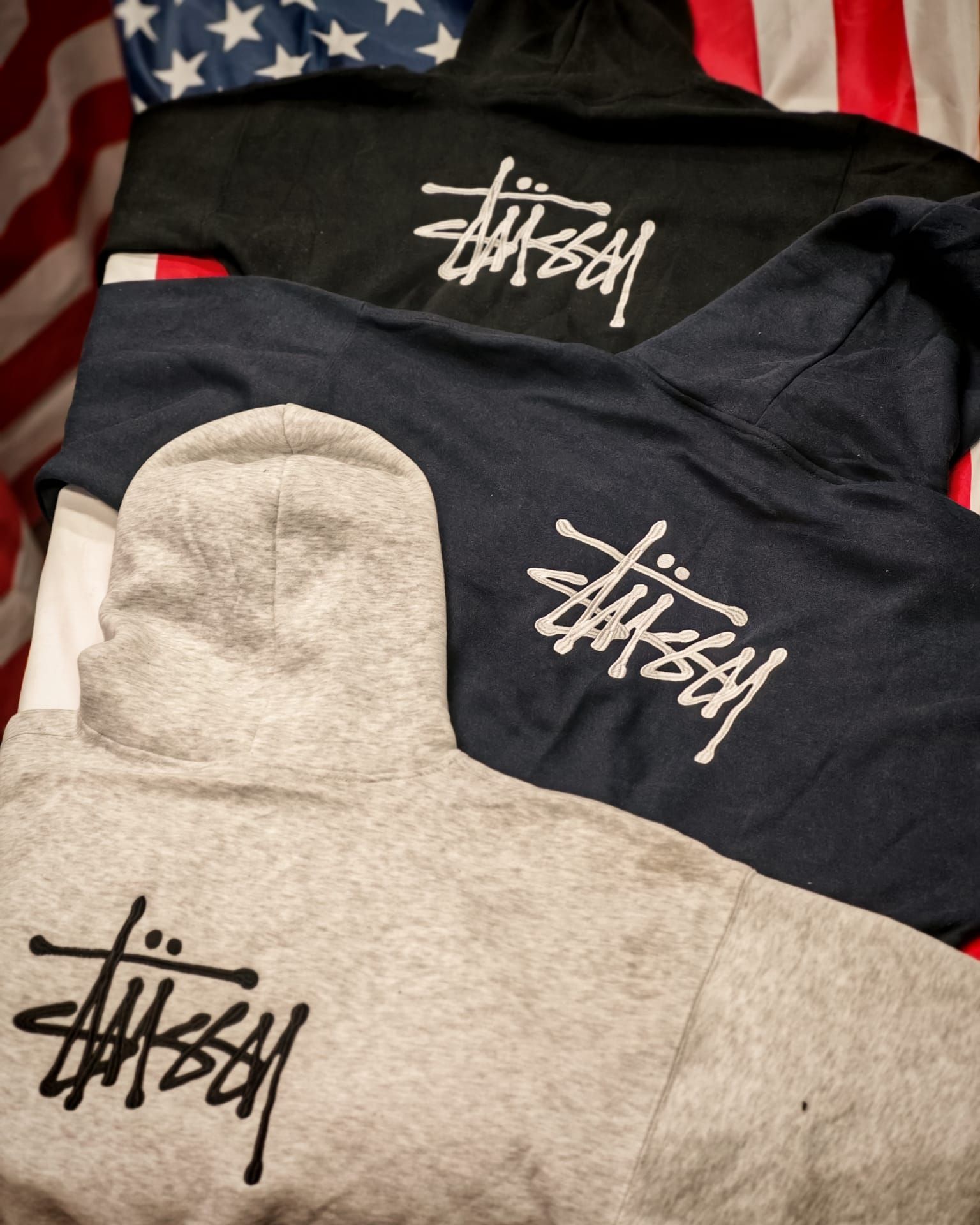 Tre felpe con cappuccio Stüssy, una nera, una blu navy e una grigio melange, impilate una sull'altra.