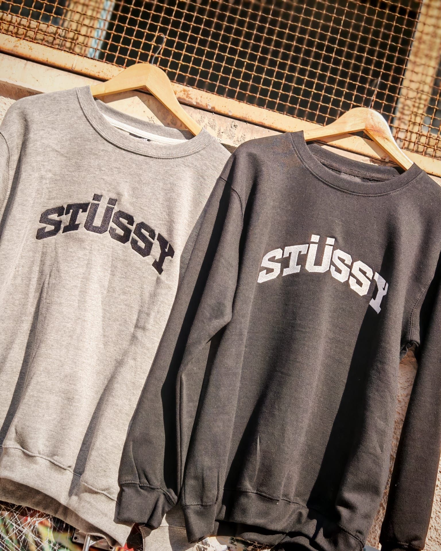 Due felpe grigie Stüssy appese a grucce di legno, esposte una accanto all'altra.