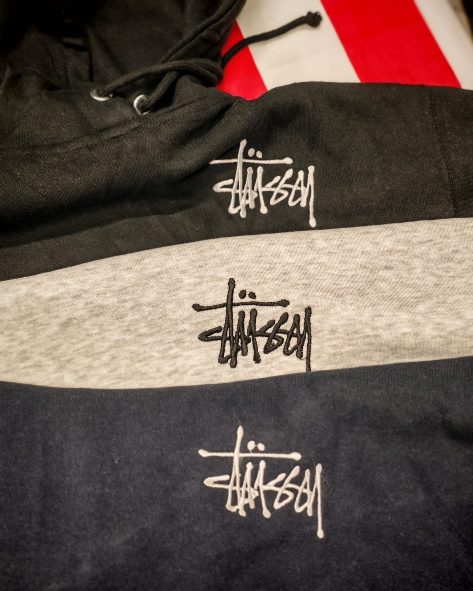 Tre felpe con cappuccio Stüssy sovrapposte, nei colori nero, grigio e blu navy, con stampa del logo sul petto.