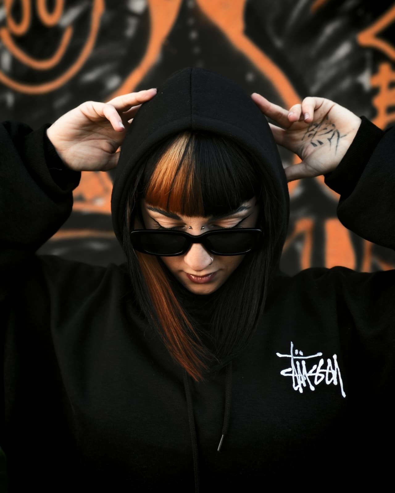 Persona con felpa nera con cappuccio e occhiali da sole che si sistema il cappuccio, con sfondo di graffiti arancioni.