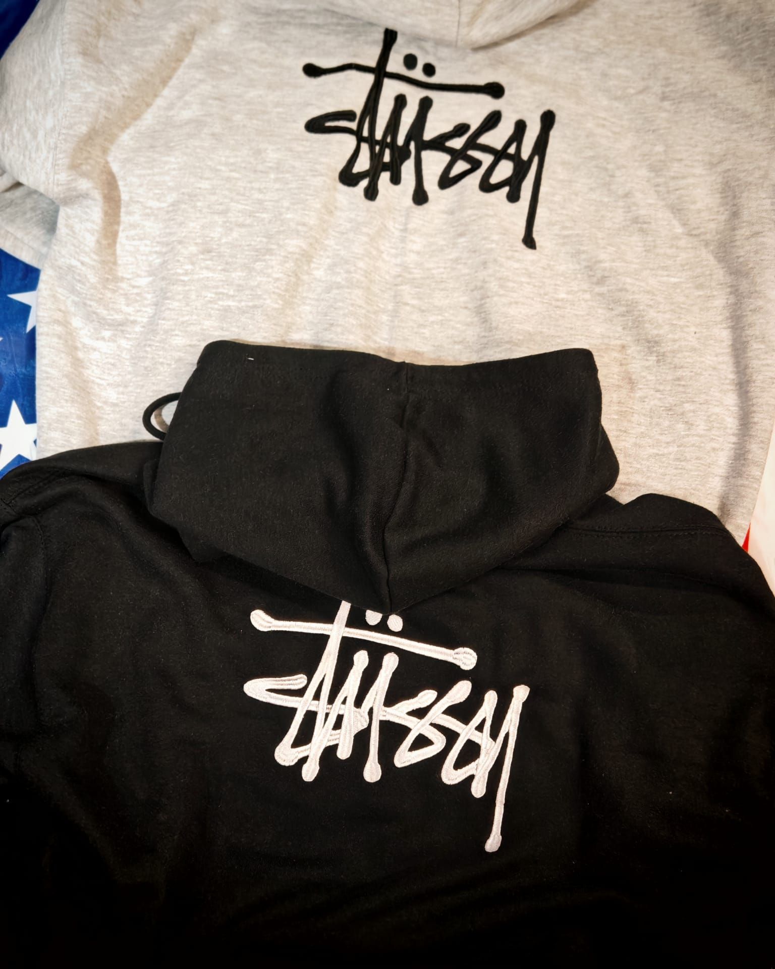 Felpe con cappuccio nere e grigie con logo Stüssy bianco, disposte una accanto all'altra su un letto.