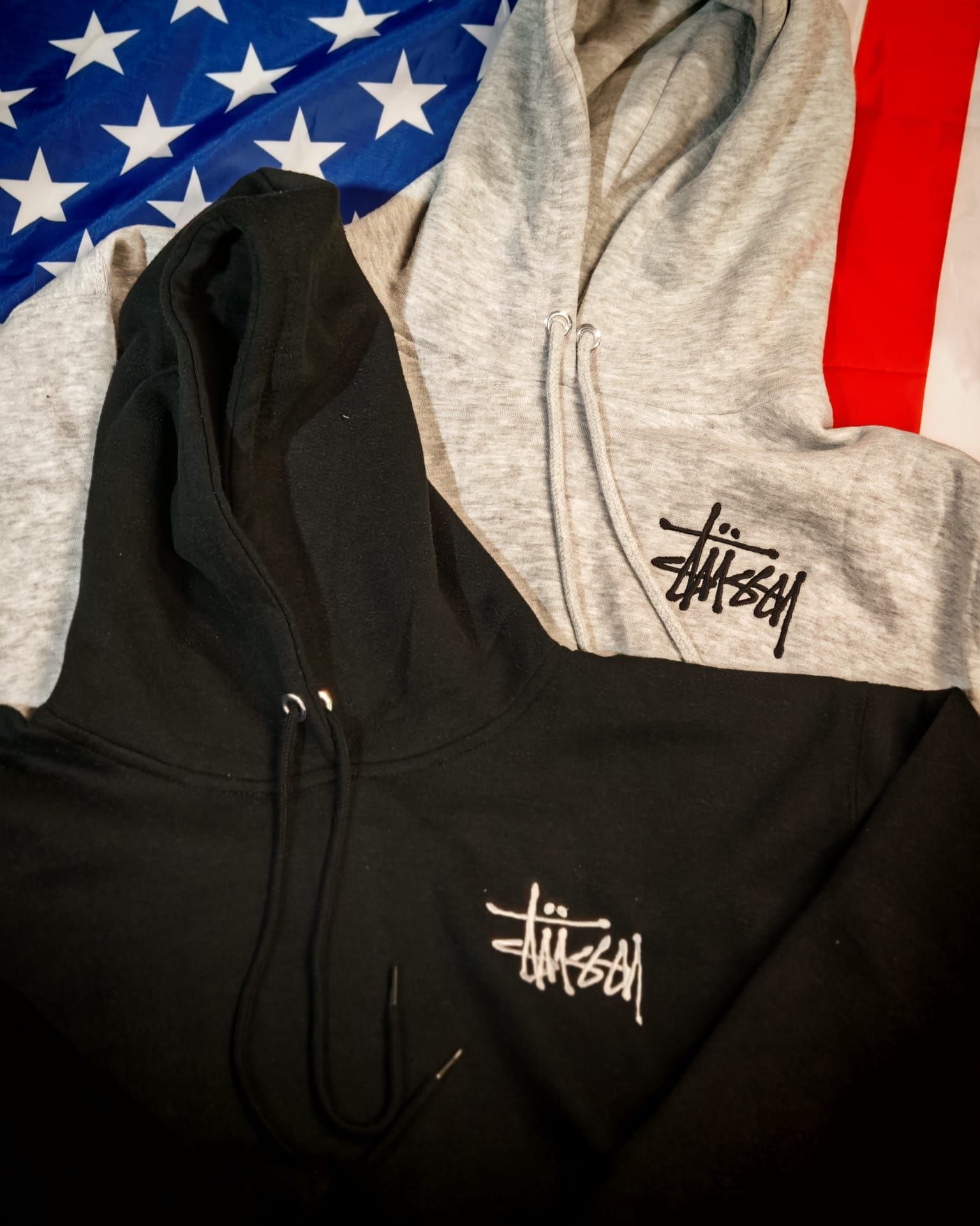 Due felpe con cappuccio, una nera e una grigia, con logo Stussy bianco su sfondo a stelle e strisce.