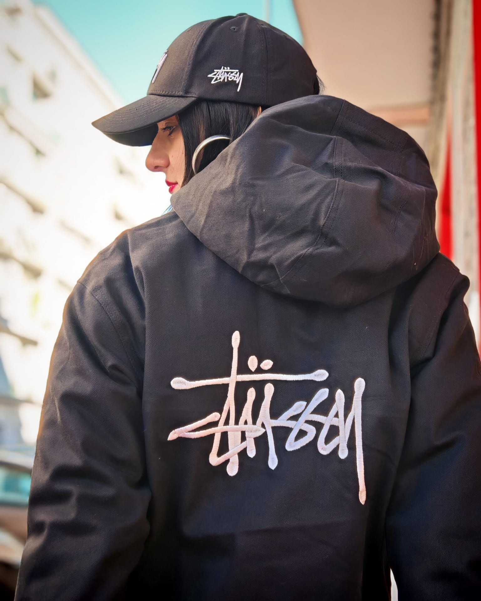 Persona con giacca e berretto neri Stüssy, vista di spalle in una strada cittadina.