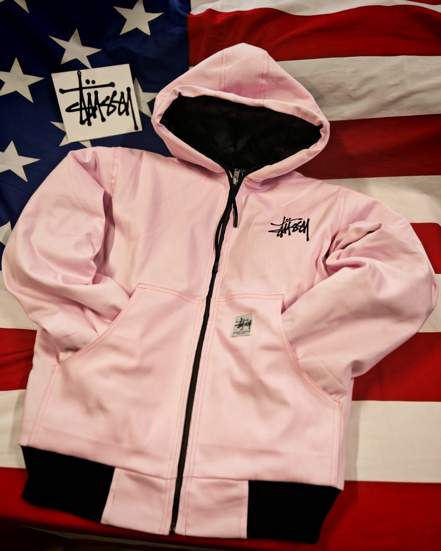 Felpa rosa con zip, finiture e loghi neri, su uno sfondo con la bandiera americana.