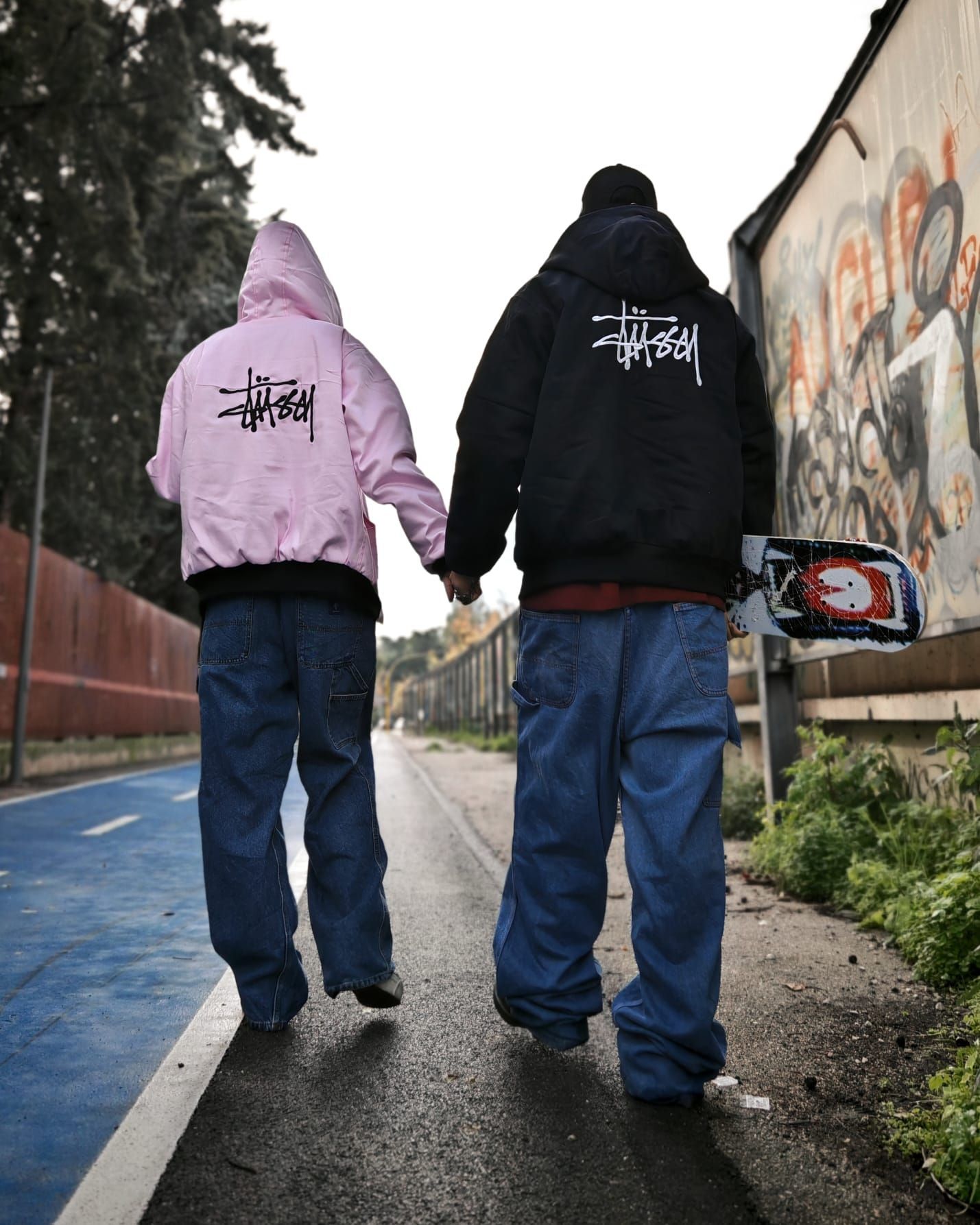 Due persone con indosso delle felpe con cappuccio si allontanano lungo una strada bagnata, costeggiando un muro ricoperto di graffiti.