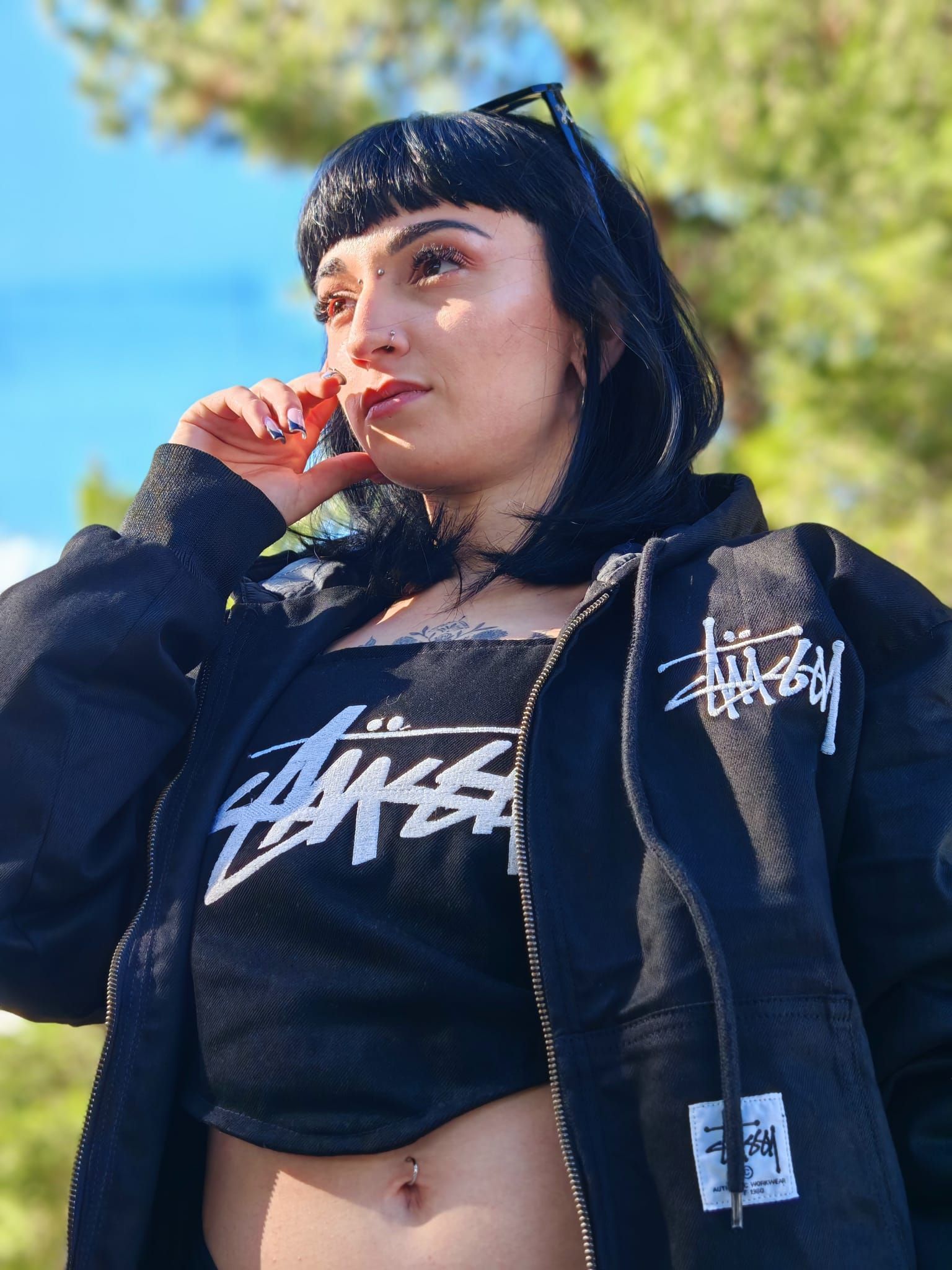 Una donna con indosso un completo nero di Stüssy posa all'aperto contro un cielo azzurro, con una mano vicino al viso.