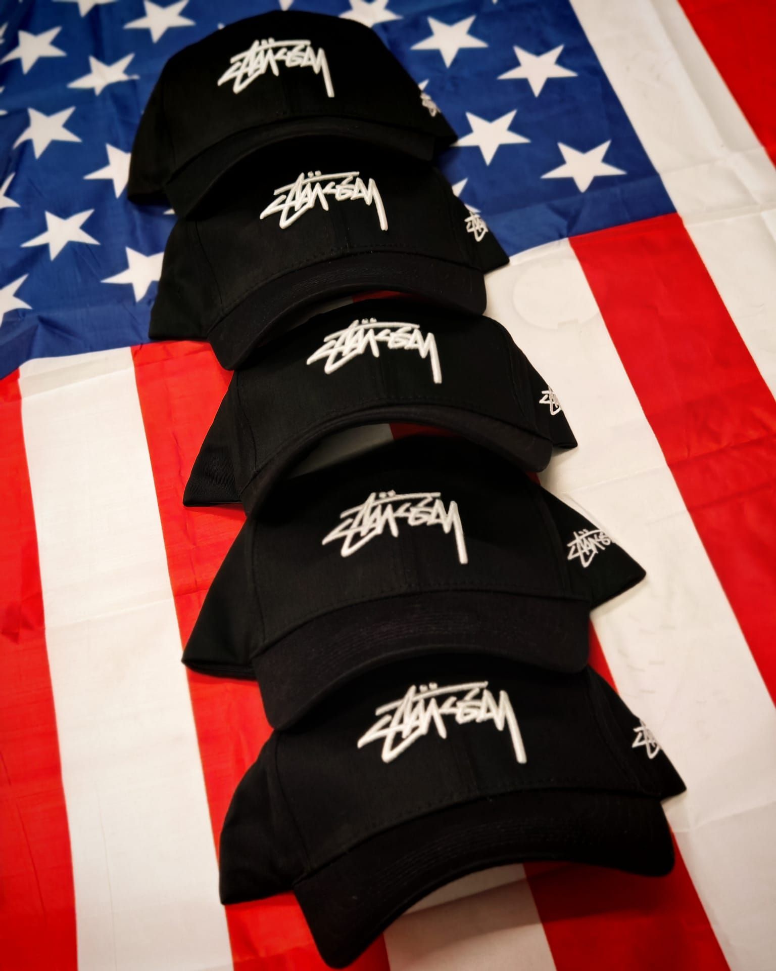 Cinque cappellini neri Stüssy impilati su uno sfondo con la bandiera americana.