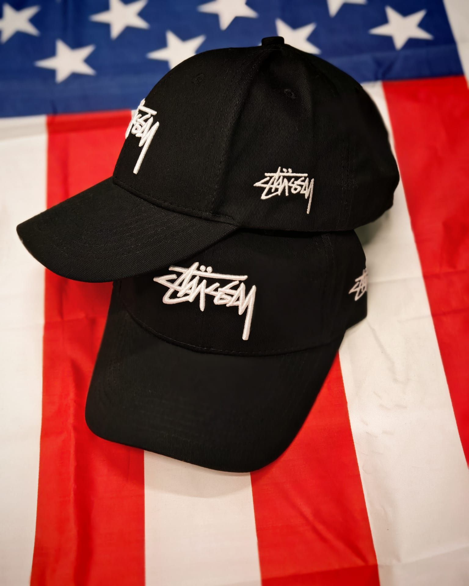 Due cappellini neri Stüssy impilati su uno sfondo con la bandiera americana.
