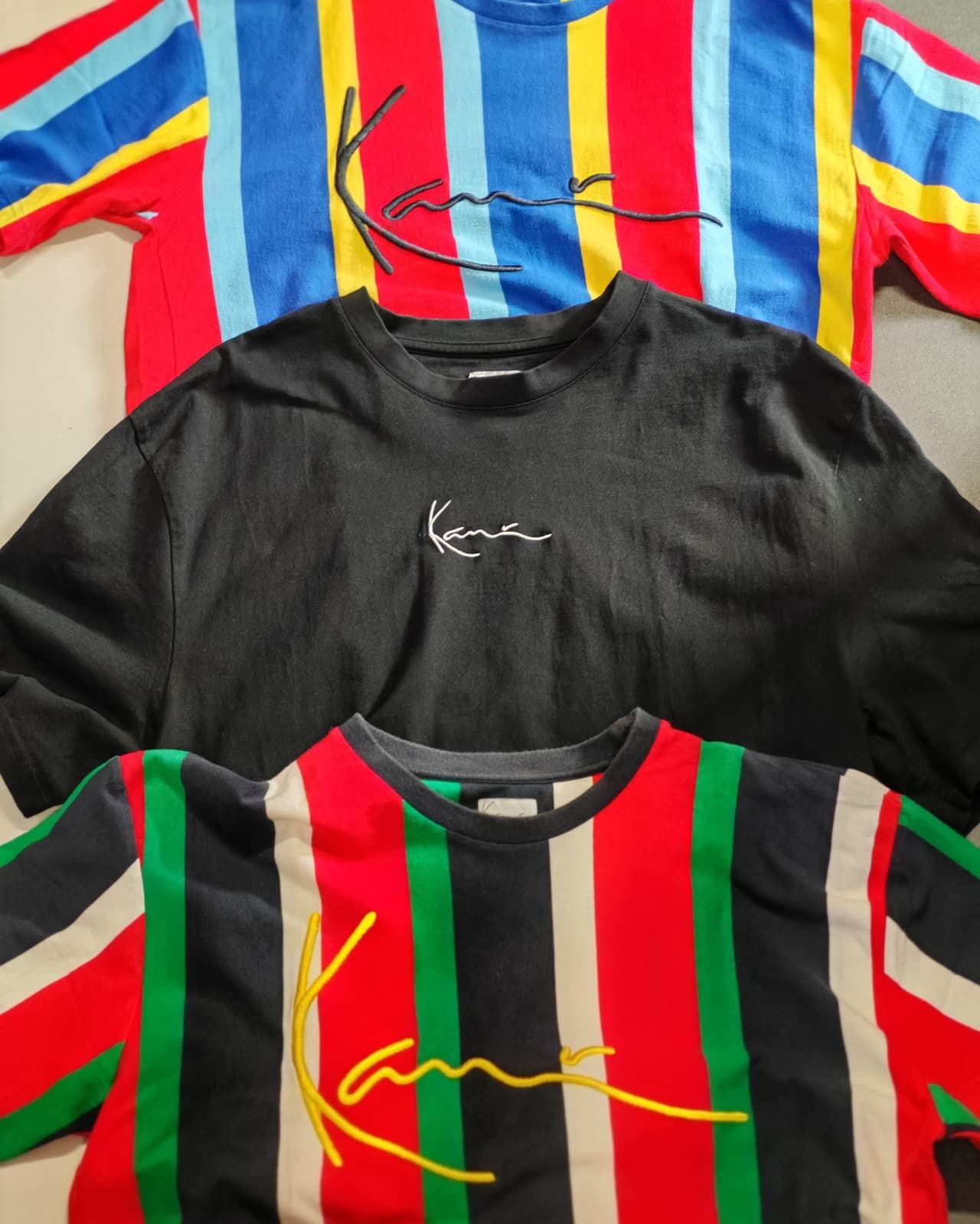 Tre magliette colorate con il marchio Kani: una nera, una a righe multicolore e una a righe rosse e verdi.