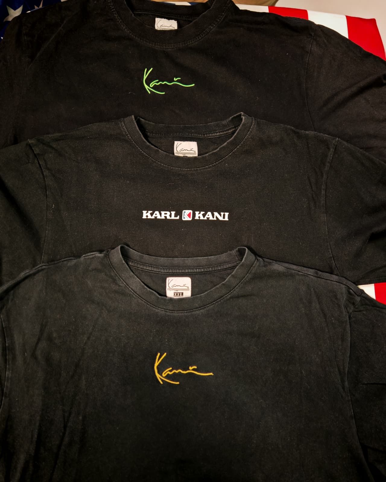 Tre t-shirt nere di Karl Kani impilate, con stampe del logo in verde, bianco e oro.