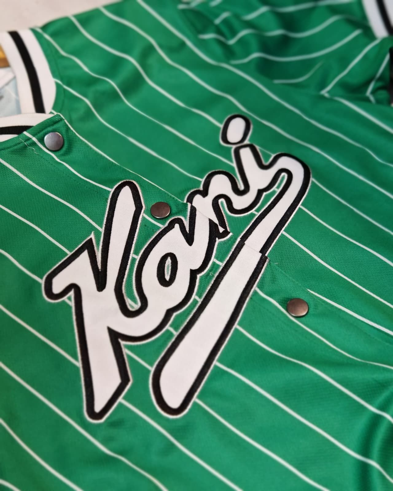 Maglia sportiva verde a righe sottili con logo in corsivo bianco e nero sul petto.