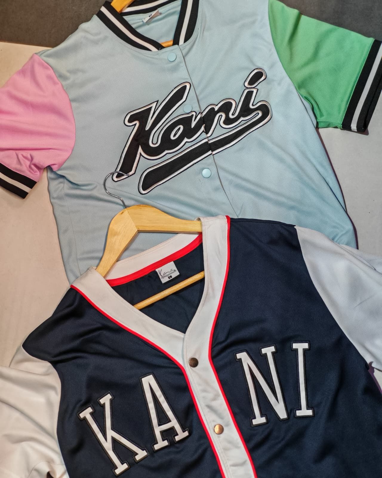 Due maglie da baseball Kani, una blu navy e grigia, l'altra multicolore pastello, distese su una superficie