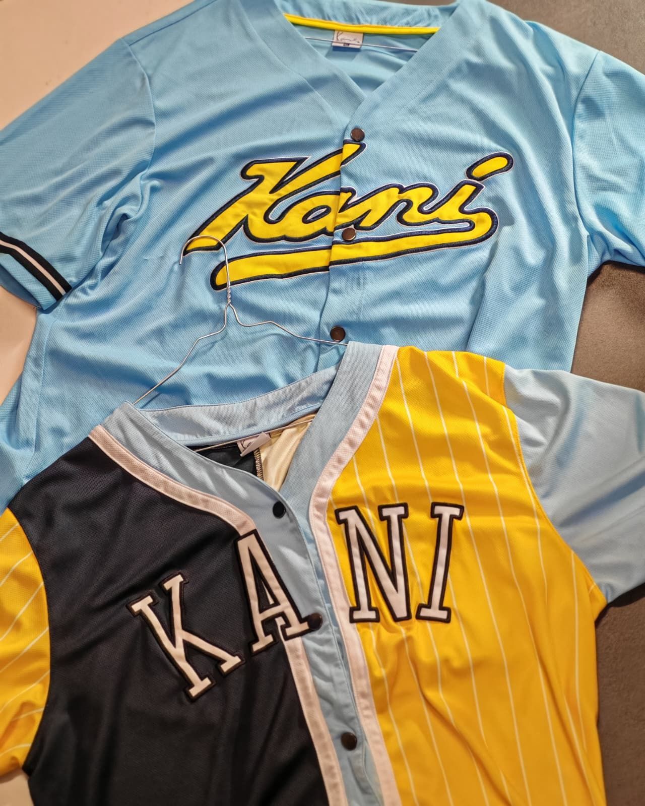 Due maglie Kani piegate insieme, una azzurra con logo giallo e una nera e gialla con scritte bianche.