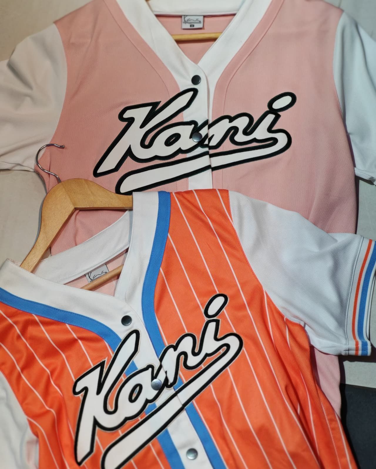Due maglie sportive Kani, una rosa e bianca e una arancione e bianca con finiture blu, appese.