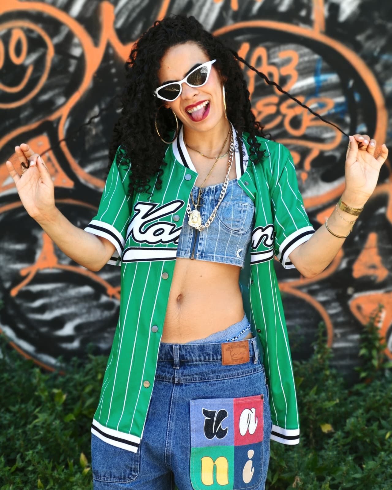 Donna con occhiali da sole in posa con le braccia alzate, indossa una maglia verde e un completo in denim davanti a un muro ricoperto di graffiti.