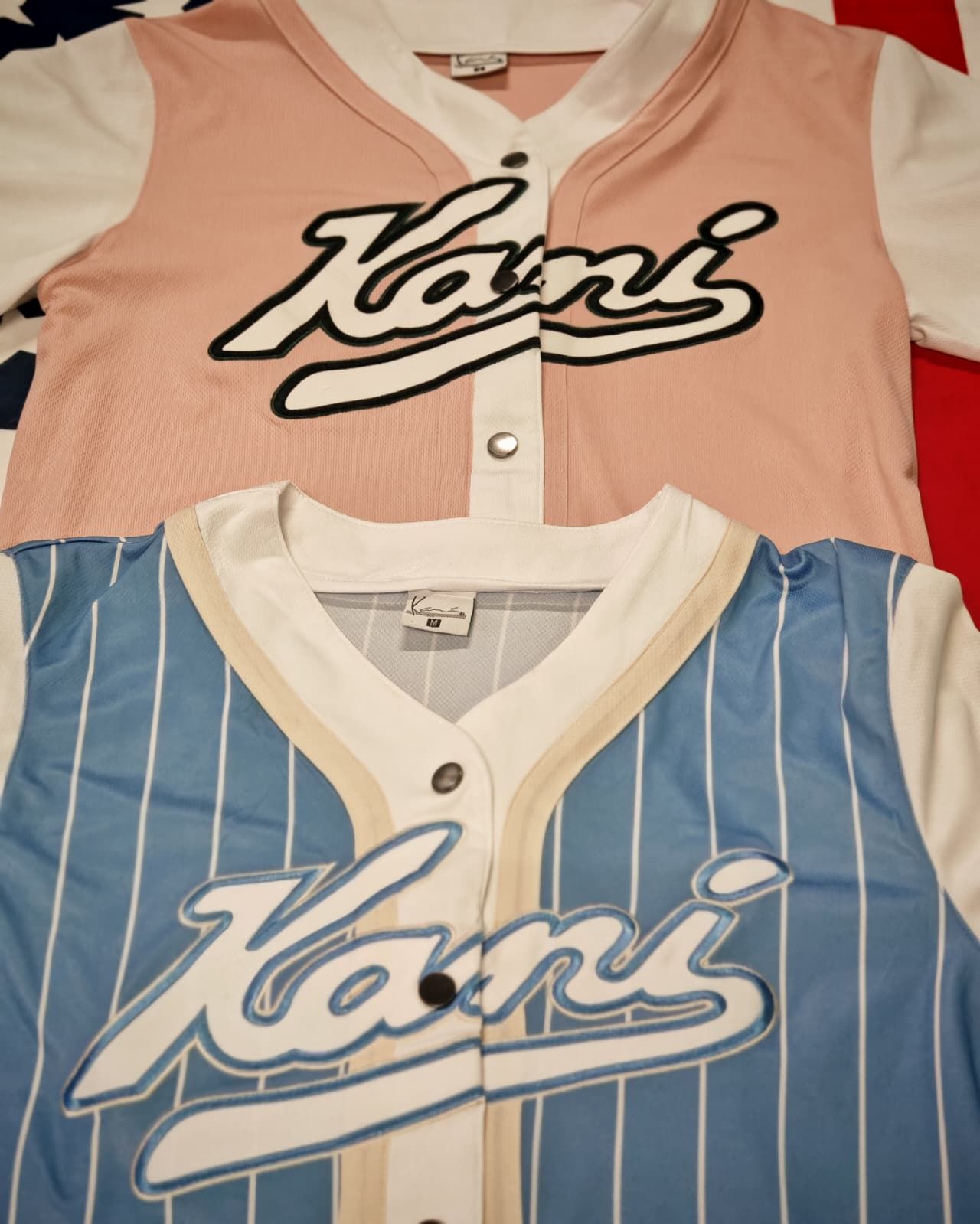 Due maglie da baseball Karmi, una rosa e una blu con righe bianche, disposte una accanto all'altra.