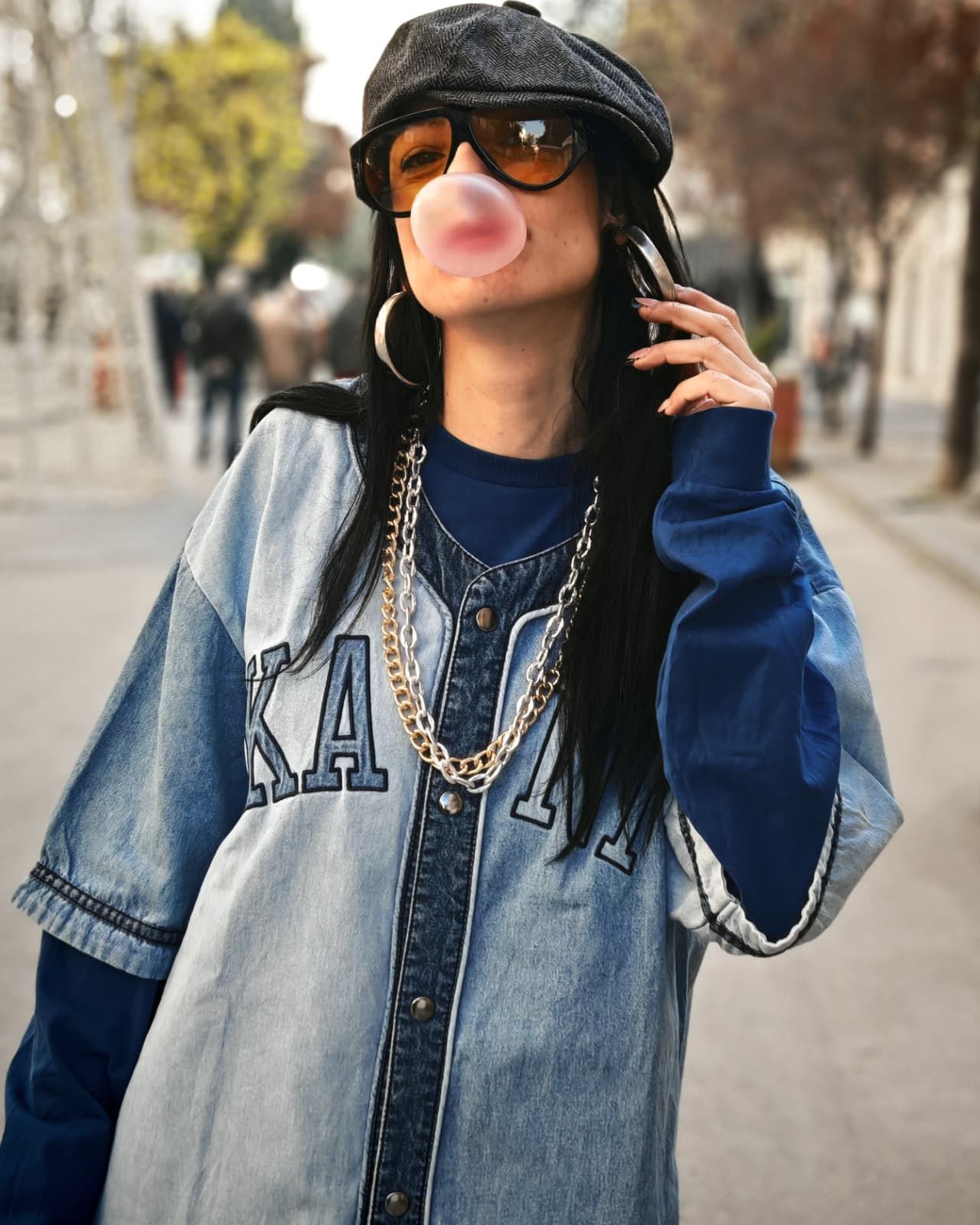 Una persona con una camicia di jeans e un berretto nero, soffia una bolla di gomma da masticare rosa su una strada cittadina.