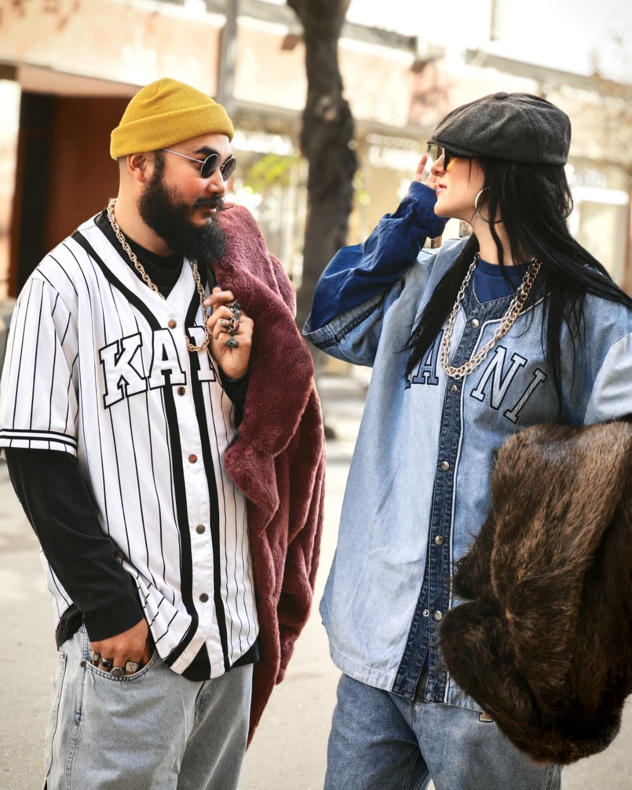 Due persone eleganti che chiacchierano all'aperto, indossando occhiali da sole, cappelli e abbigliamento streetwear a strati.