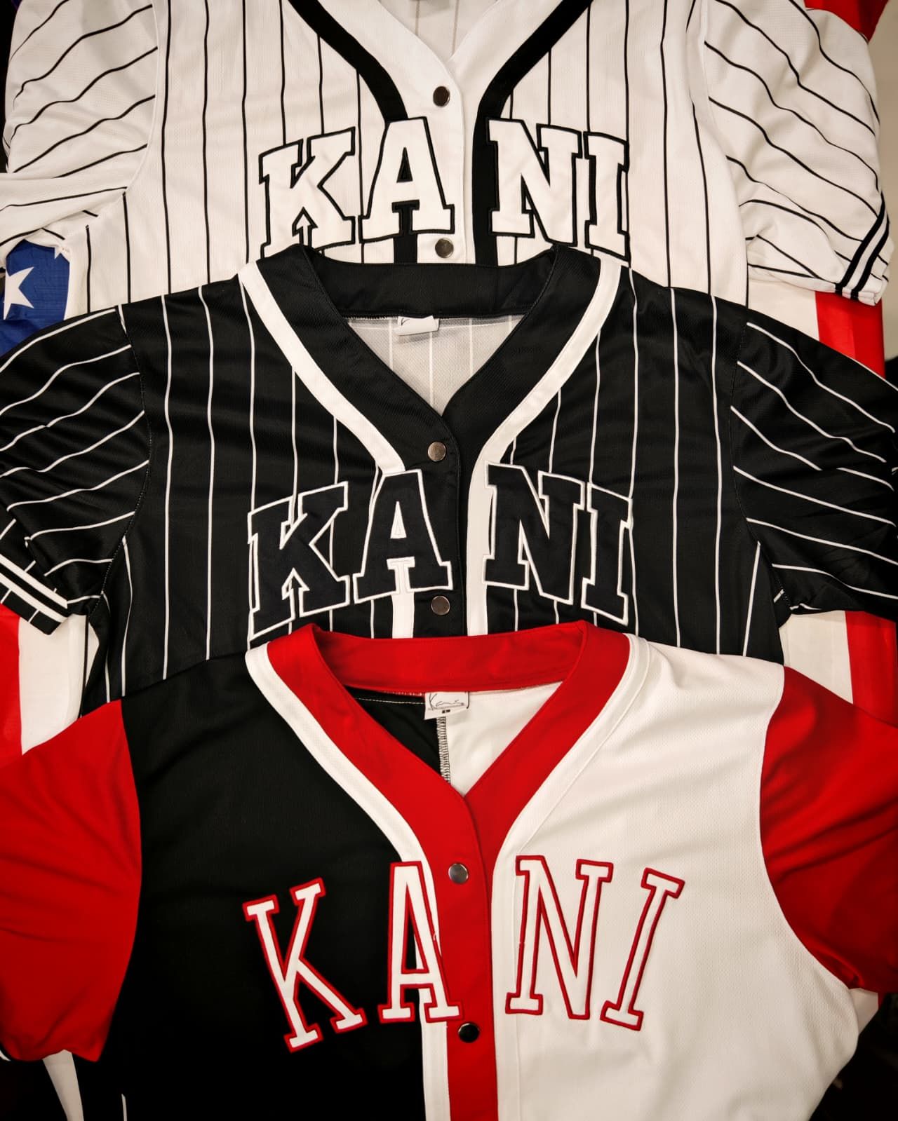 Pila di tre maglie KANI nei colori bianco, nero e rosso con scritte in grassetto