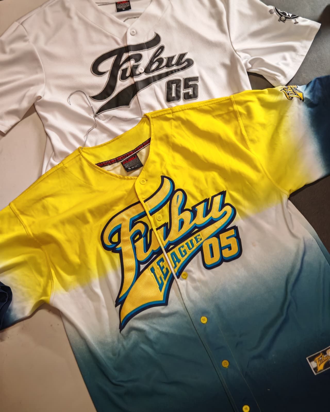 Due maglie da baseball Fubu, bianca e con sfumatura dal giallo al blu, disposte in piano con un grande logo e la scritta