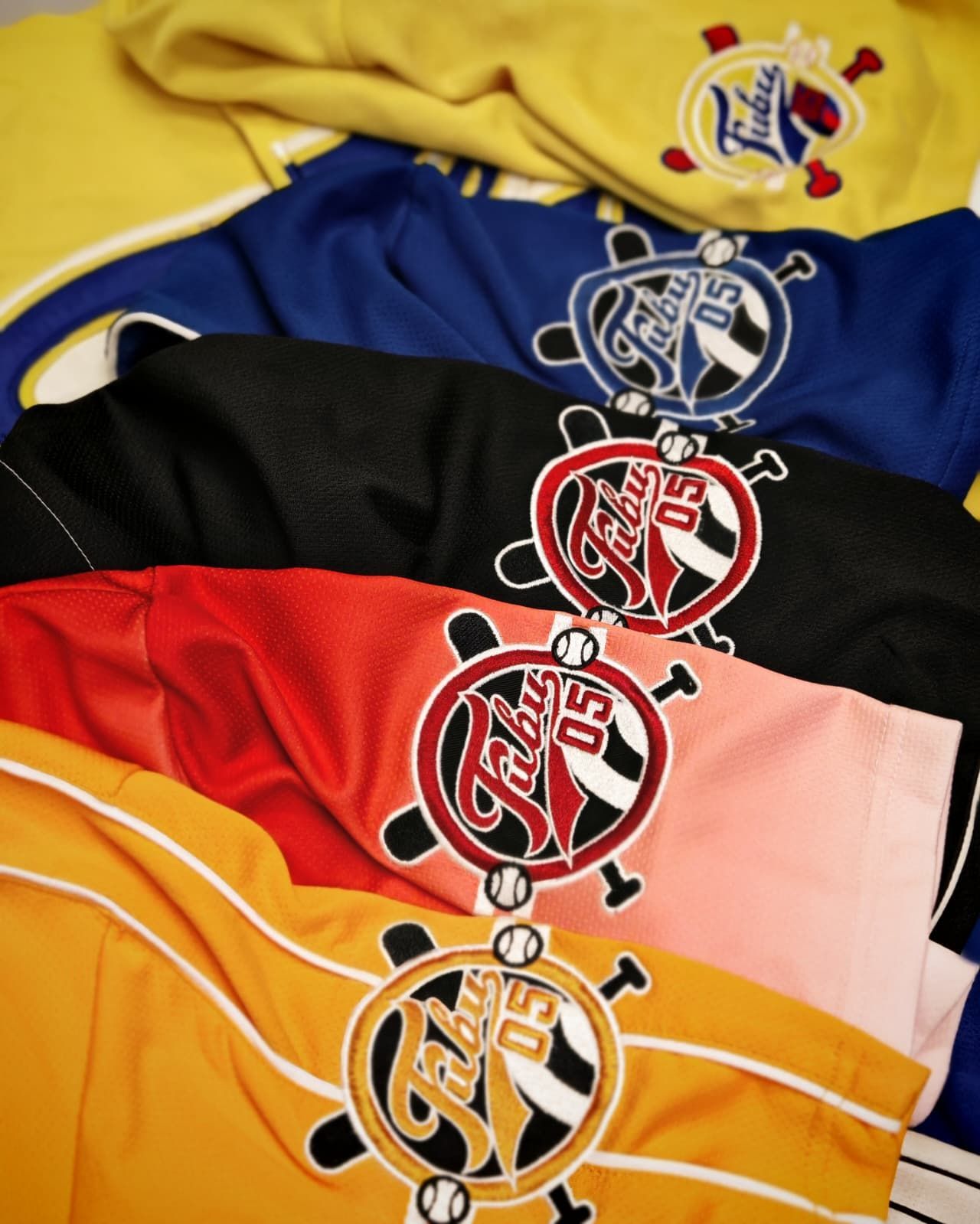 Maglie sportive colorate impilate, con loghi circolari nei colori giallo, blu, nero, rosso, rosa e arancione.