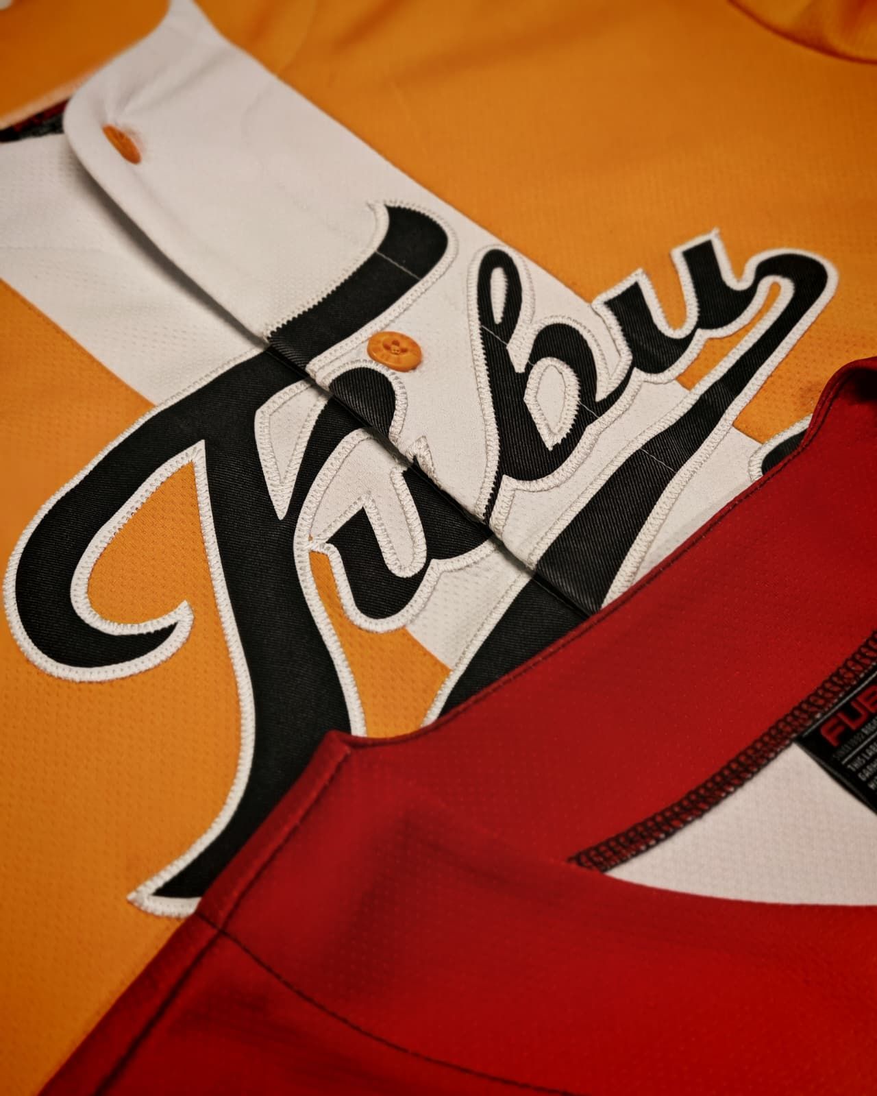Maglie sportive arancioni e bianche con logo in corsivo nero e un indumento rosso in primo piano.