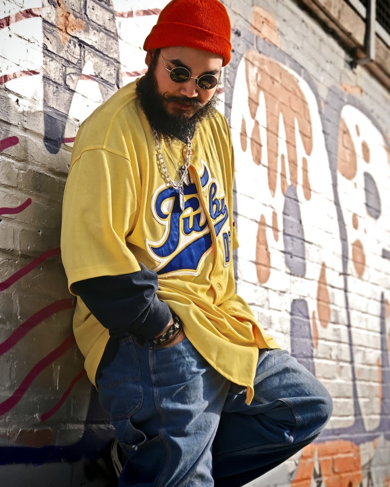Un uomo appoggiato a un muro di mattoni indossa una maglia gialla, un berretto rosso, occhiali da sole e jeans blu.