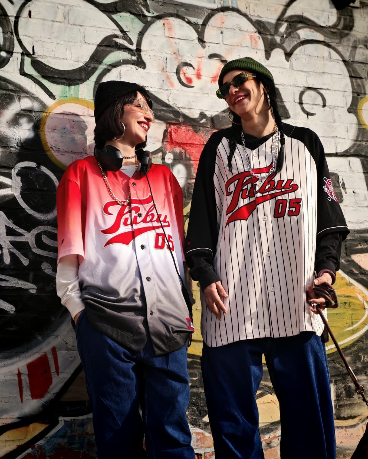 Due persone con indosso maglie da baseball ridono davanti a un muro ricoperto di graffiti.
