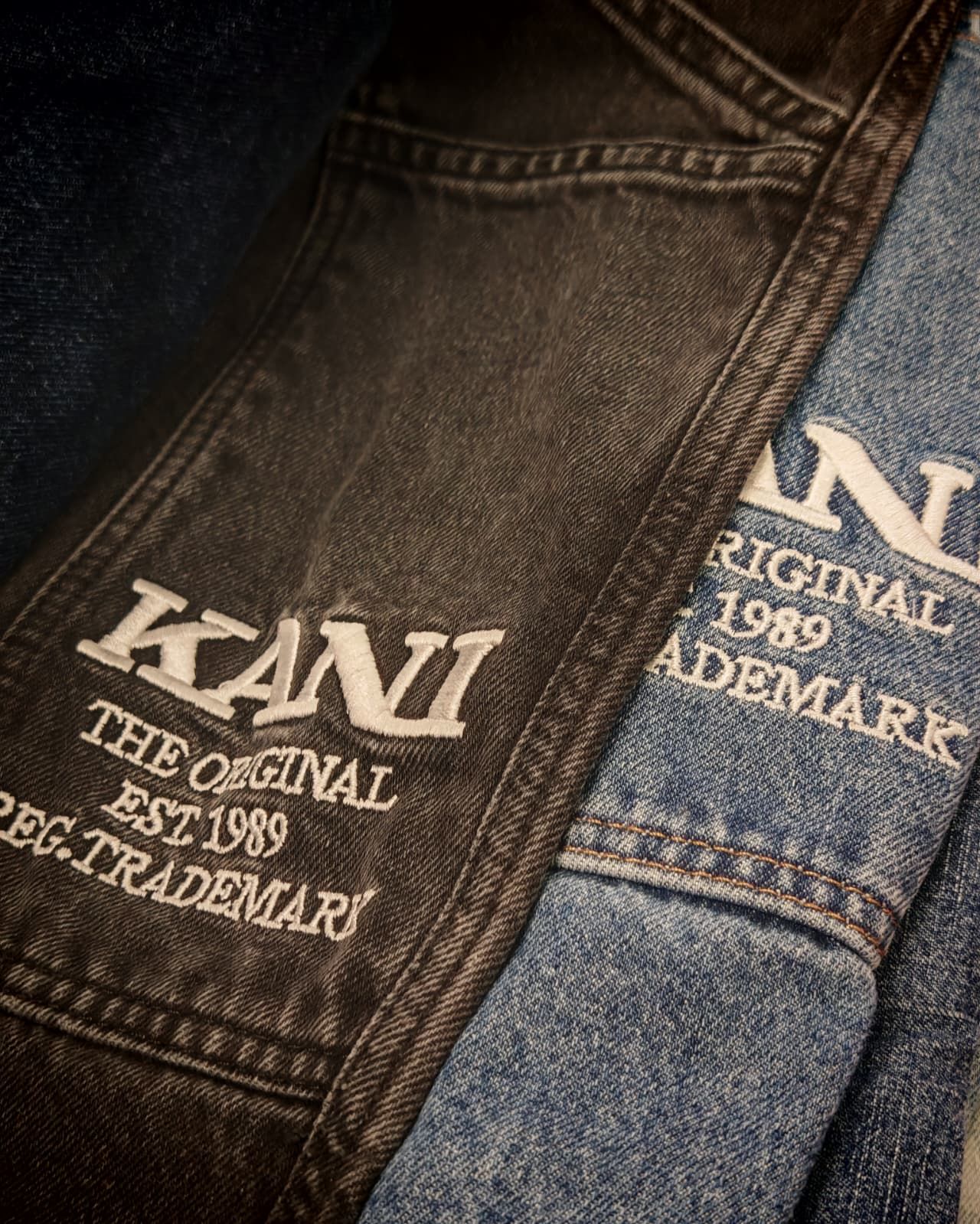 Primo piano di due paia di jeans con il marchio KANI ben visibile su denim marrone scuro e blu.