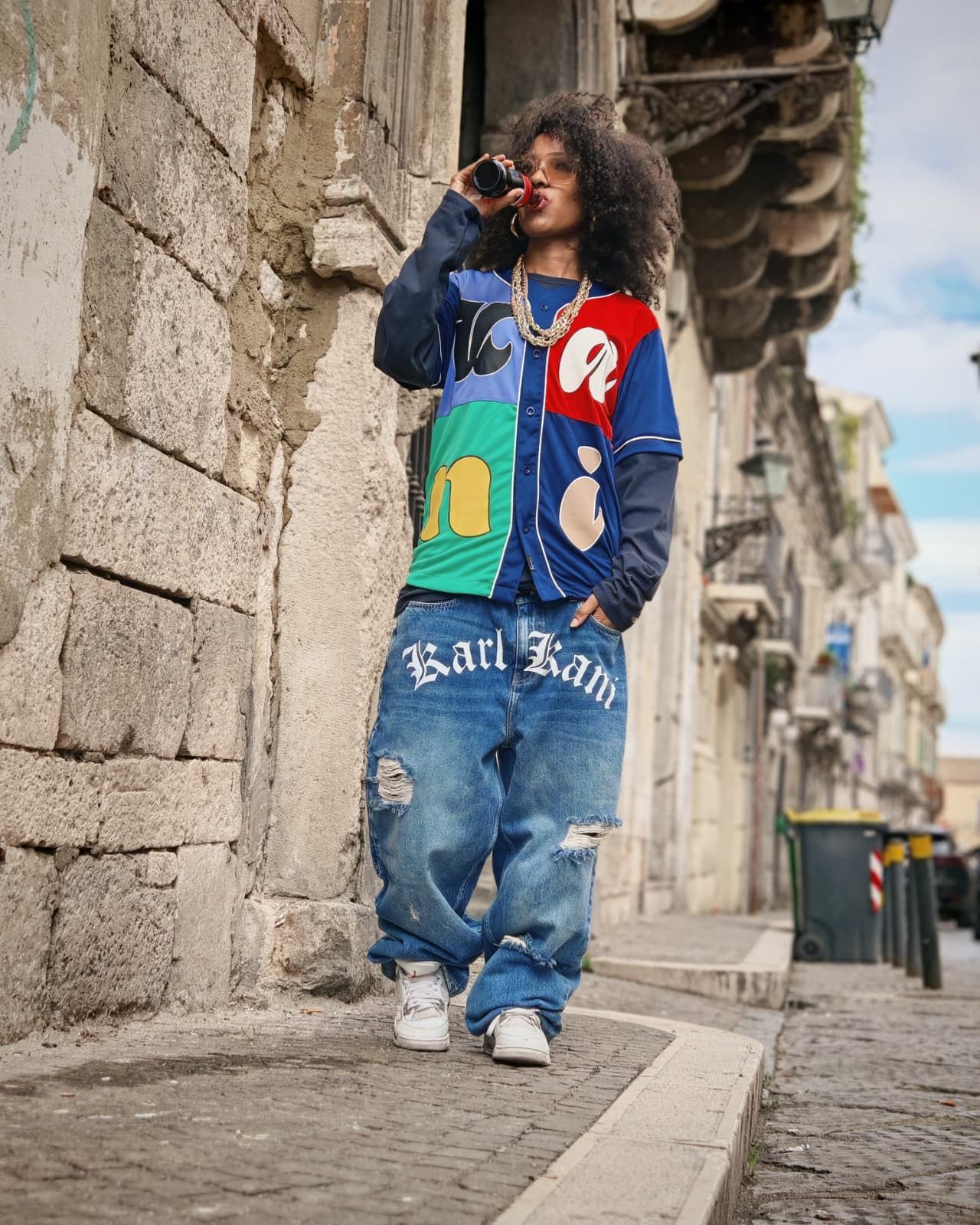 Persona con indosso abiti streetwear colorati in piedi accanto a un muro di pietra su una strada acciottolata.