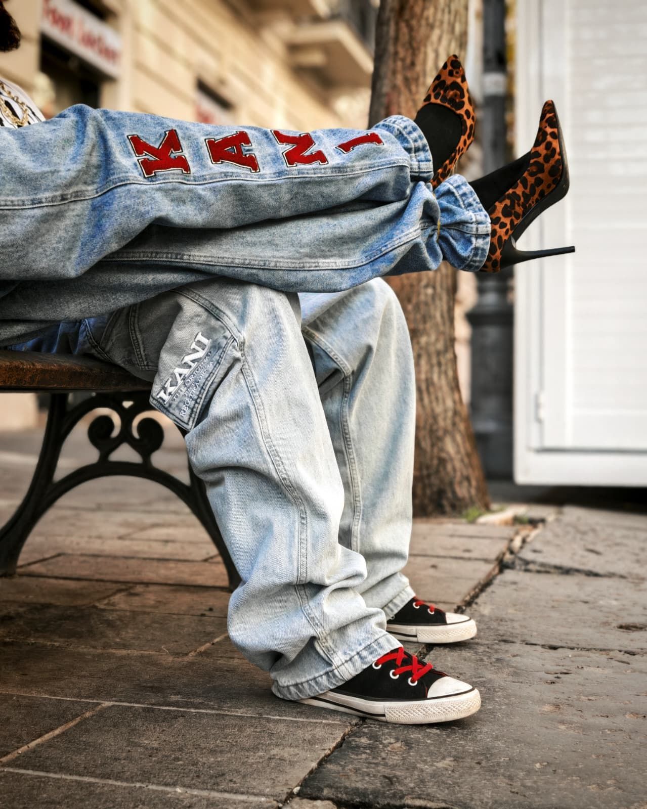 Una persona è seduta su una panchina con le gambe incrociate, indossa jeans larghi e chiari e scarpe da ginnastica nere con dettagli rossi.