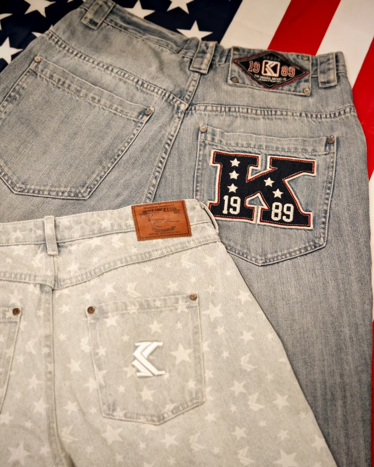 Due paia di jeans con toppe ricamate, uno grigio chiaro e uno bianco, adagiati su uno sfondo con la bandiera americana.